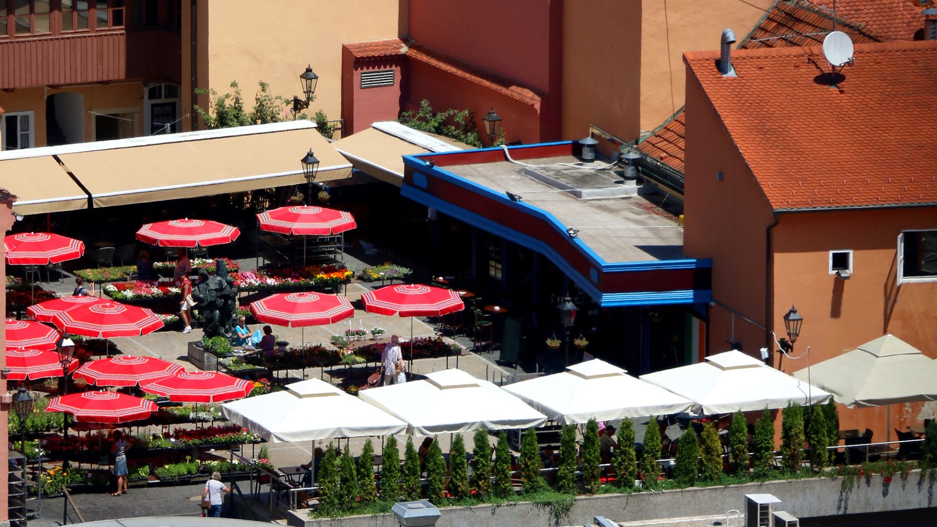 An aerial view of the bustling Dolac Market in Zagreb, Croatia, featuring numerous stalls with vibrant red umbrellas covering fresh produce and goods, surrounded by buildings with traditional architecture. 