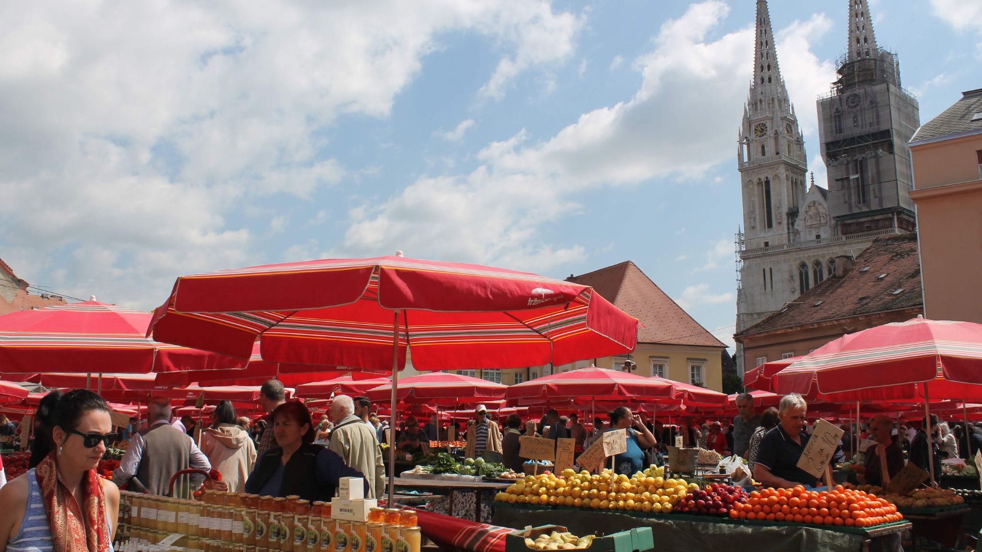 A bustling open-air farmers' market in Zagreb, Croatia, with numerous red-striped umbrellas sheltering stalls laden with fresh produce like fruits and vegetables, and various local goods. In the background, the spires of Zagreb Cathedral are visible under a partly cloudy sky.