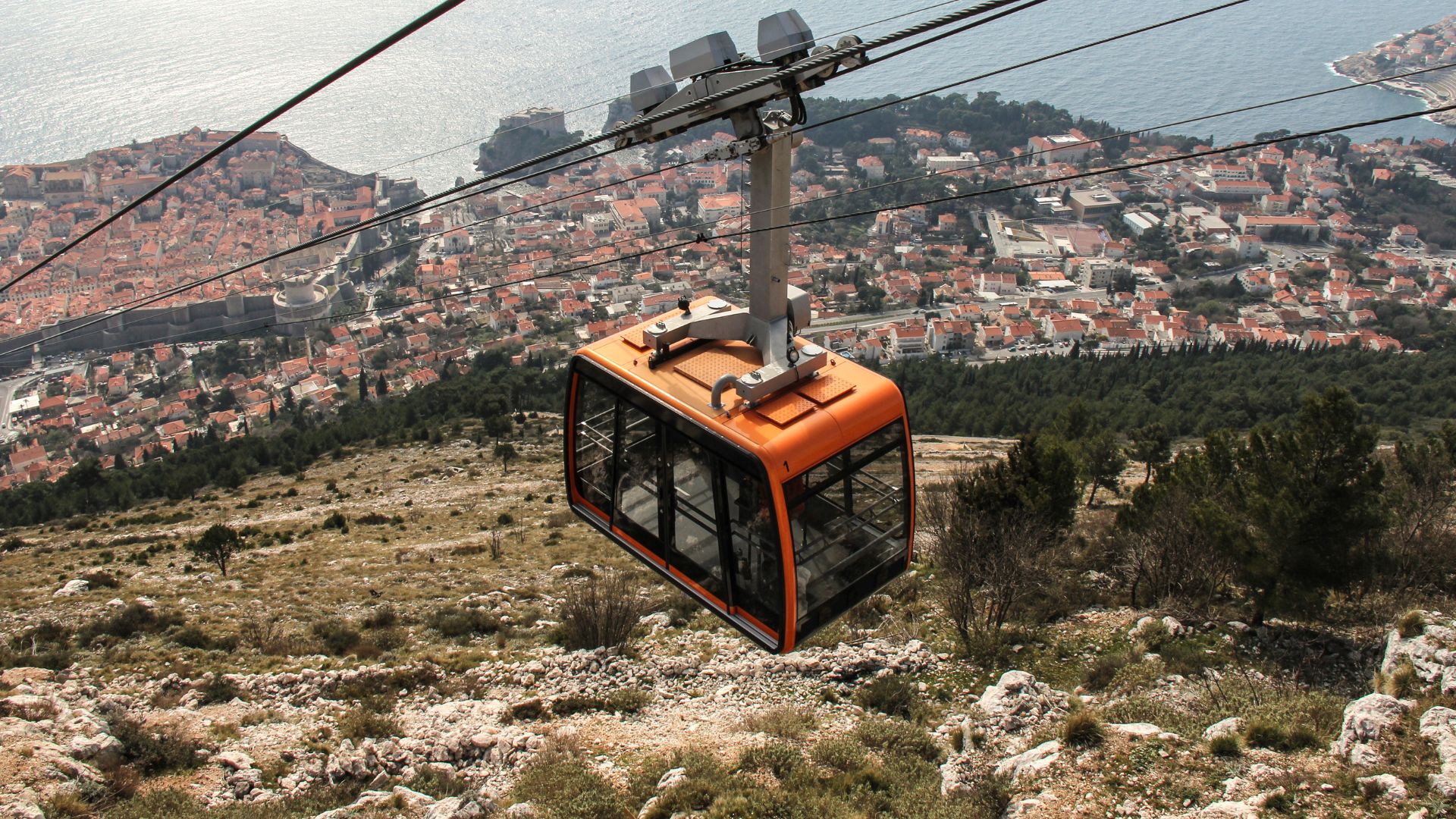 An orange cable car gondola ascends a rocky, sparsely vegetated mountainside, with the historic, red-roofed city of Dubrovnik and the blue Adriatic Sea visible in the background under a hazy sky.