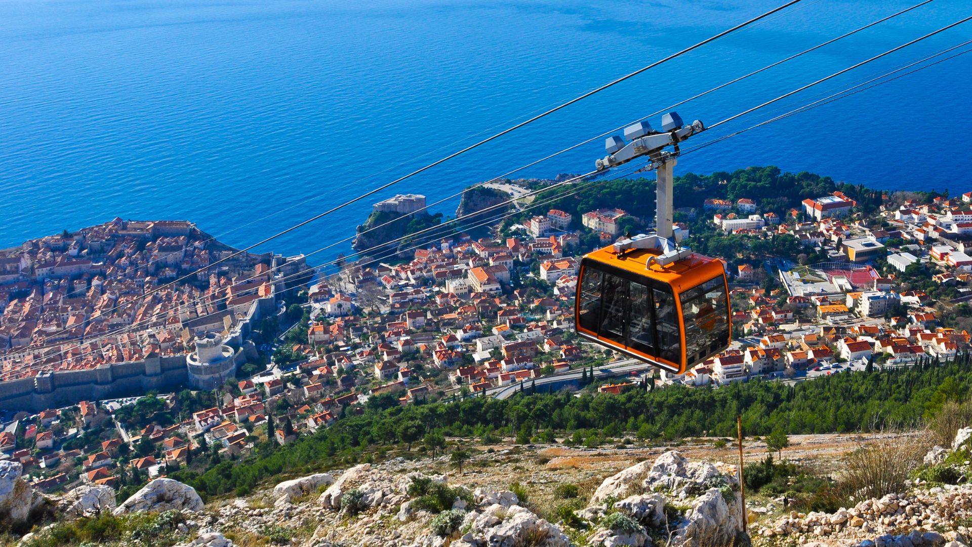 An orange cable car cabin ascends a mountain, with the historic walled city of Dubrovnik and the vast blue Adriatic Sea visible below under a clear sky.
