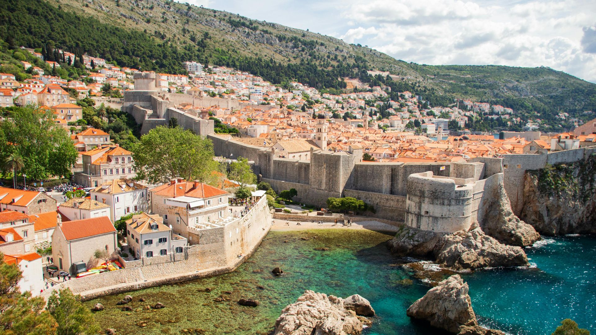 A vibrant, elevated view of Dubrovnik's Old Town, a historic walled city with orange-tiled roofs nestled against a lush green mountain and overlooking the clear turquoise waters of the Adriatic Sea, with ancient stone walls and fortifications prominent along the coastline.