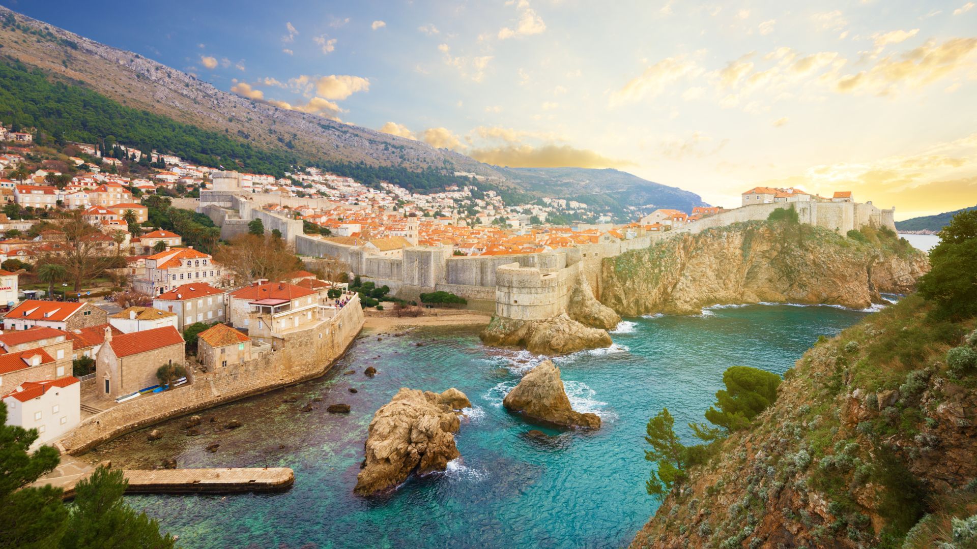 A vibrant, elevated view of Dubrovnik's Old Town, a historic walled city with orange-tiled roofs nestled against a lush green mountain and overlooking the clear turquoise waters of the Adriatic Sea, with ancient stone walls and fortifications prominent along the coastline.
