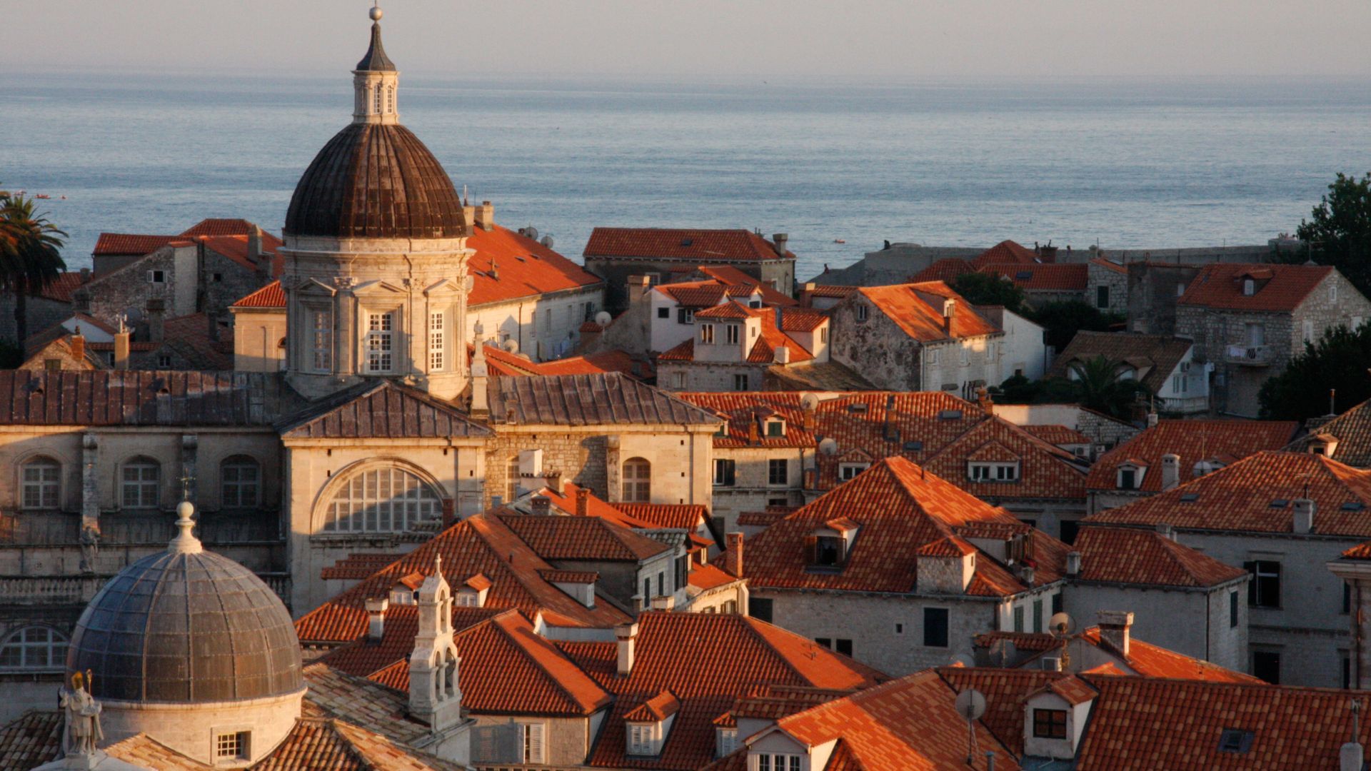  An elevated view of Dubrovnik, Croatia, showcasing its historic terracotta rooftops and a large domed cathedral, with the calm Adriatic Sea visible in the background under a soft light. 