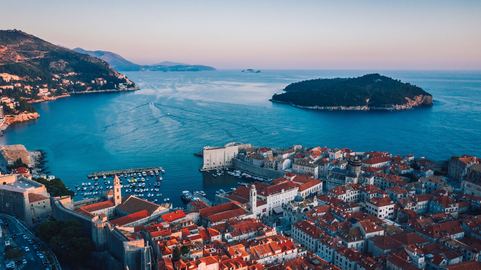 An aerial view of the historic walled city of Dubrovnik, Croatia, with its terracotta rooftops and ancient city walls, nestled along the clear blue waters of the Adriatic Sea, with a lush green island in the distance under a clear sky.