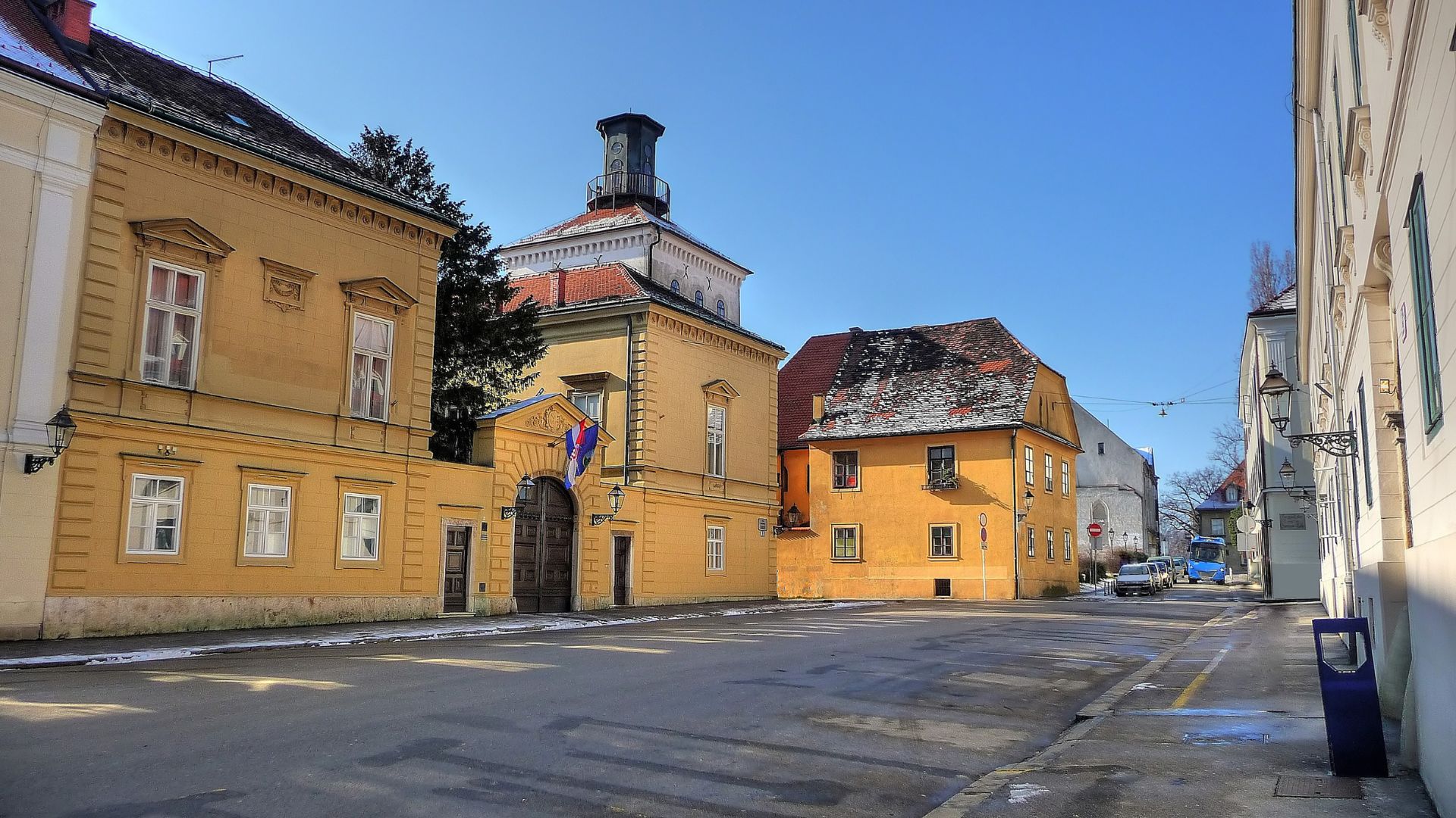  A sunny street scene in Zagreb's Upper Town, featuring the yellow-hued Dverce Palace with its distinctive tower, flanked by other historic buildings, with a clear blue sky overhead and a hint of snow on the ground.