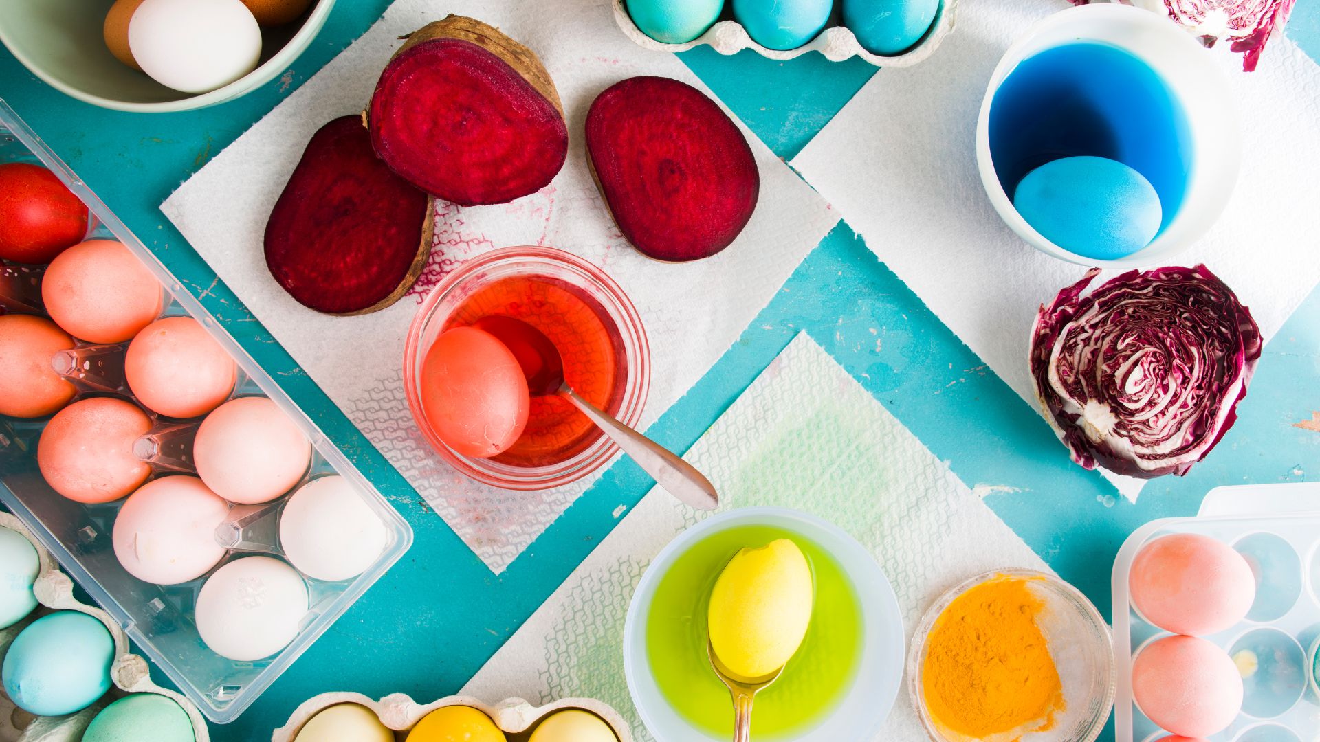 A top-down view of various natural ingredients like beets and red cabbage prepared for dyeing Easter eggs, alongside naturally dyed eggs in shades of pink, yellow, and blue in bowls and egg cartons.
