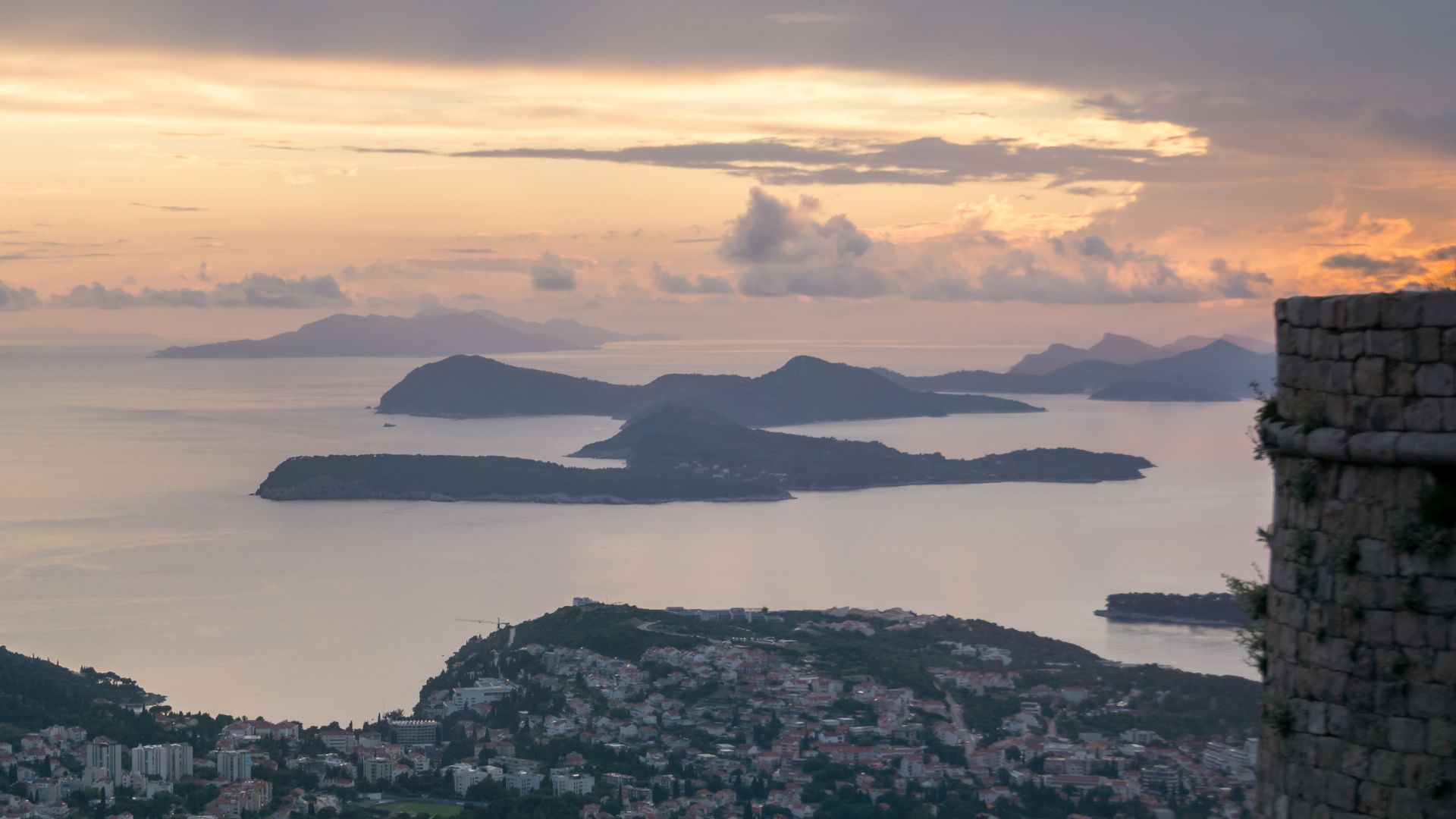 A scenic view of the Adriatic Sea at sunset, featuring several islands in the distance under a cloudy sky, with a stone fortress wall in the foreground on the right.