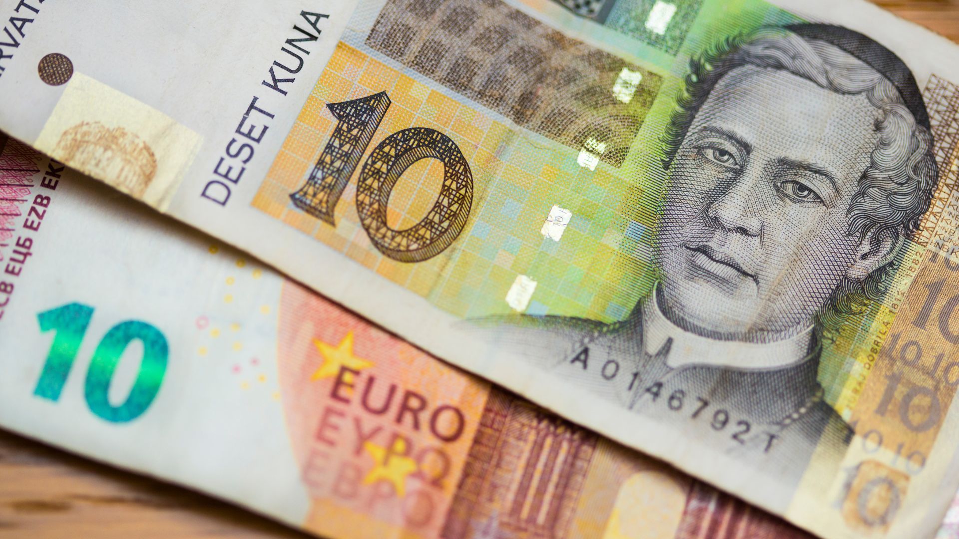 A close-up shot of two banknotes, a 10 Euro note at the bottom left and a 10 Croatian Kuna note partially overlapping it on the top right, featuring a portrait and the text "DESET KUNA."