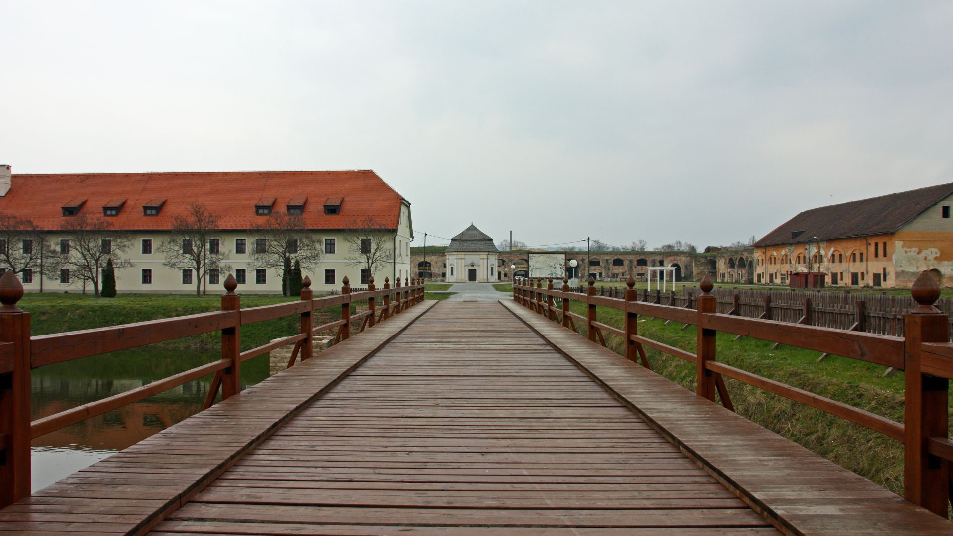 A wooden bridge extends over water towards a large, historic fortress complex with red-roofed buildings under an overcast sky.