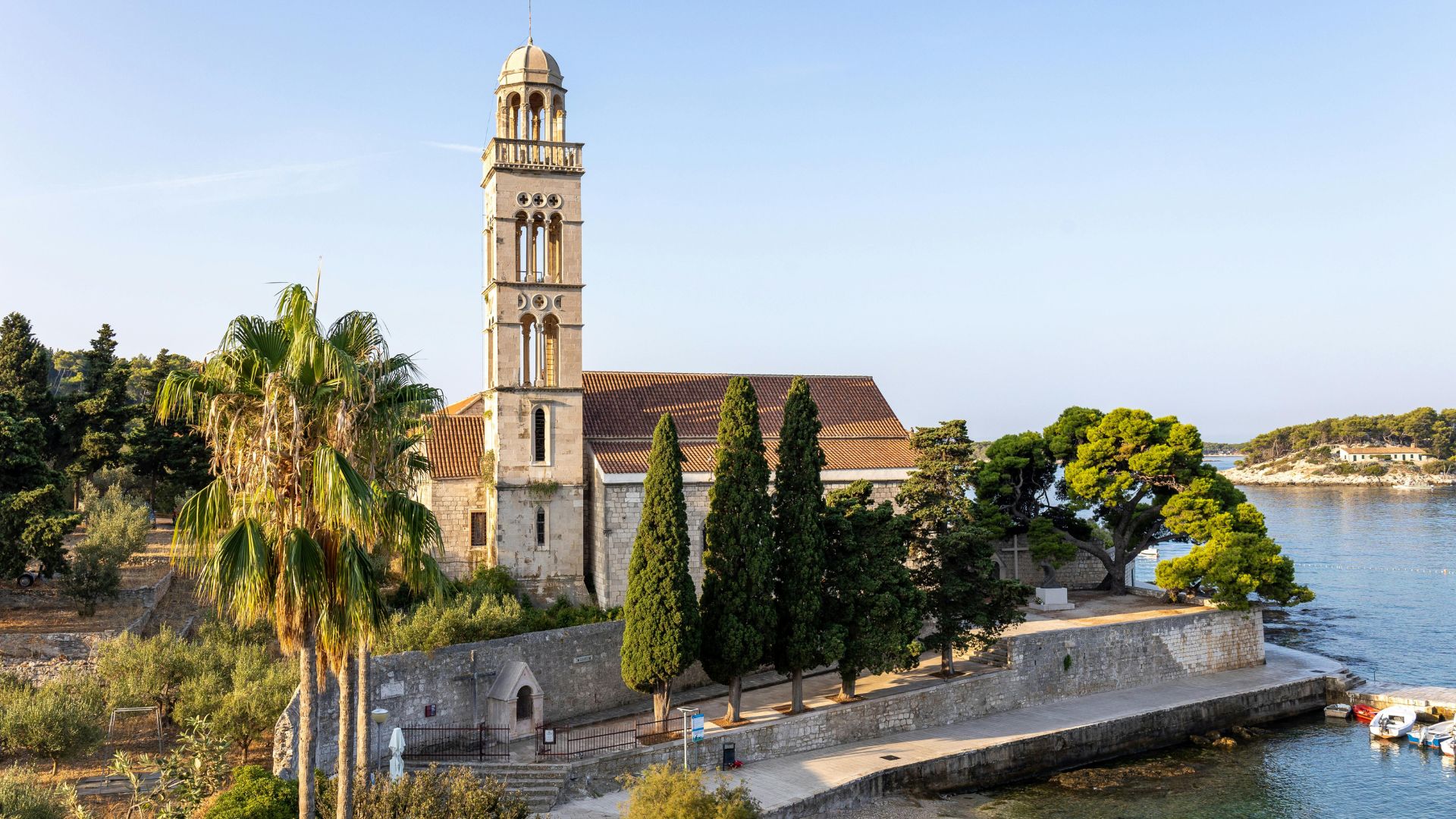 A picturesque view of the Franciscan Monastery and its bell tower, situated on the waterfront of Hvar Island, Croatia, surrounded by lush green trees and calm blue waters with small boats docked nearby.