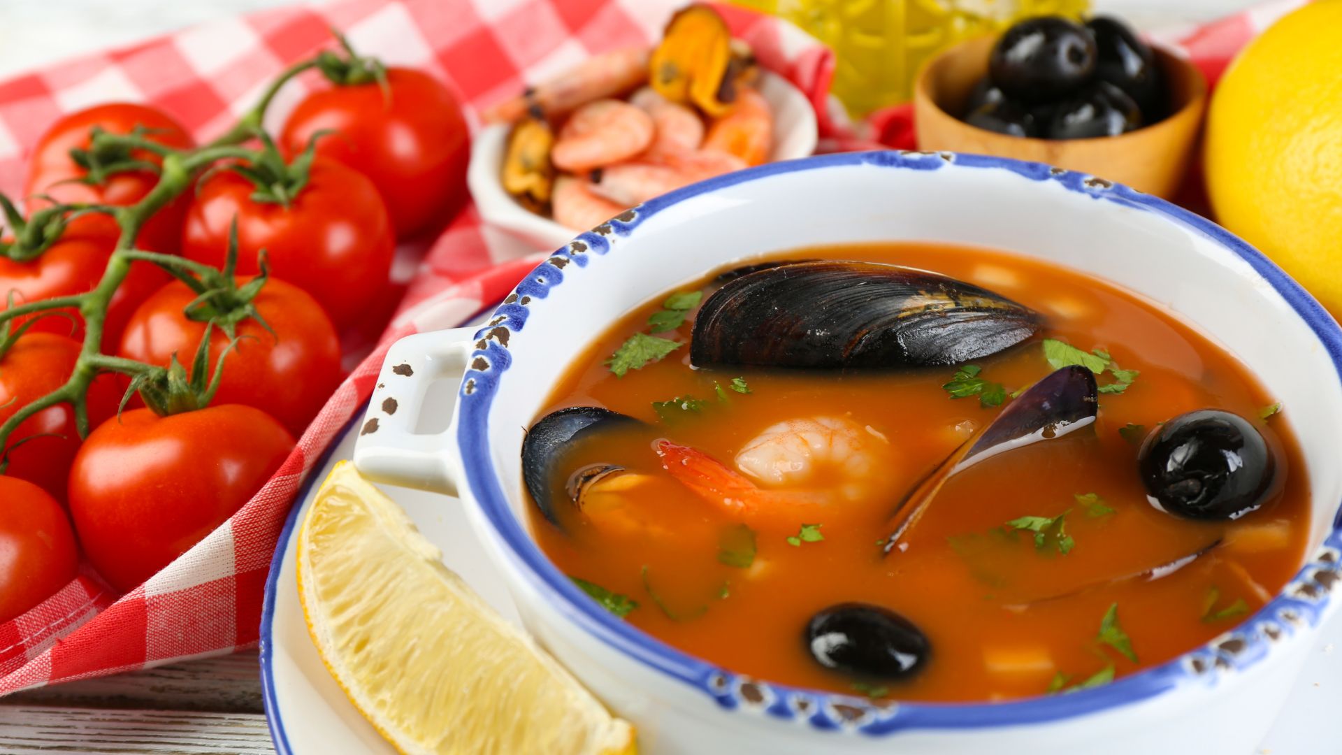 A close-up of a bowl of rich red seafood soup or stew, containing mussels, shrimp, and black olives, garnished with a lemon wedge and fresh tomatoes in the background.