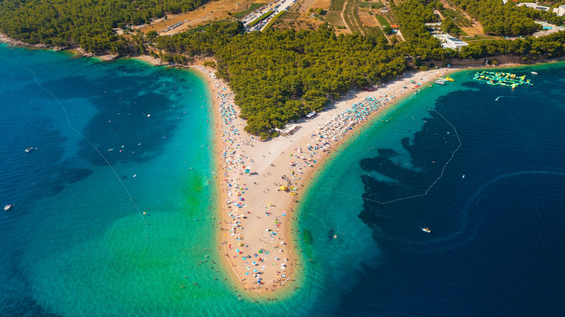 An aerial view of Zlatni Rat beach, a distinctive V-shaped pebble beach with a forested area extending into the turquoise and deep blue waters of the Adriatic Sea, populated with beachgoers and a few boats.