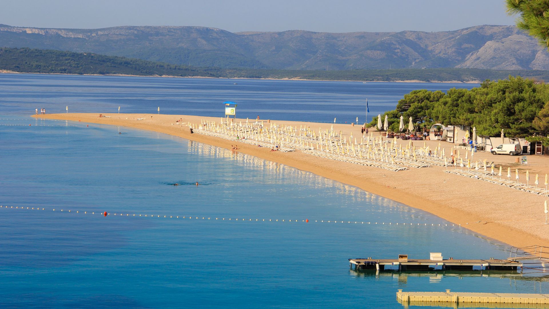 A panoramic view of Zlatni Rat beach in Croatia, showcasing its unique golden horn shape extending into the clear blue Adriatic Sea, with sunbathers and umbrellas on the pebble beach, and lush green trees bordering the shore, all set against a backdrop of distant mountains.