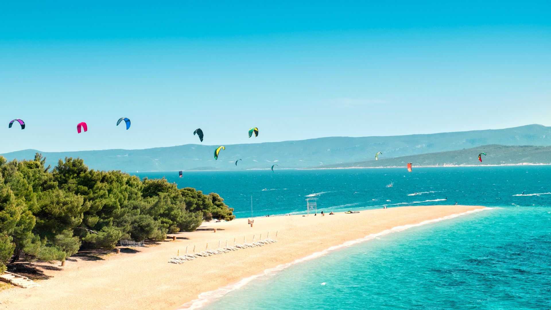 An aerial view shows the unique, golden-pebble Zlatni Rat beach, a prominent cape extending into the turquoise Adriatic Sea, lined with green pine trees, with several colorful kitesurfers enjoying the clear waters and a distant mountainous coastline under a bright blue sky.