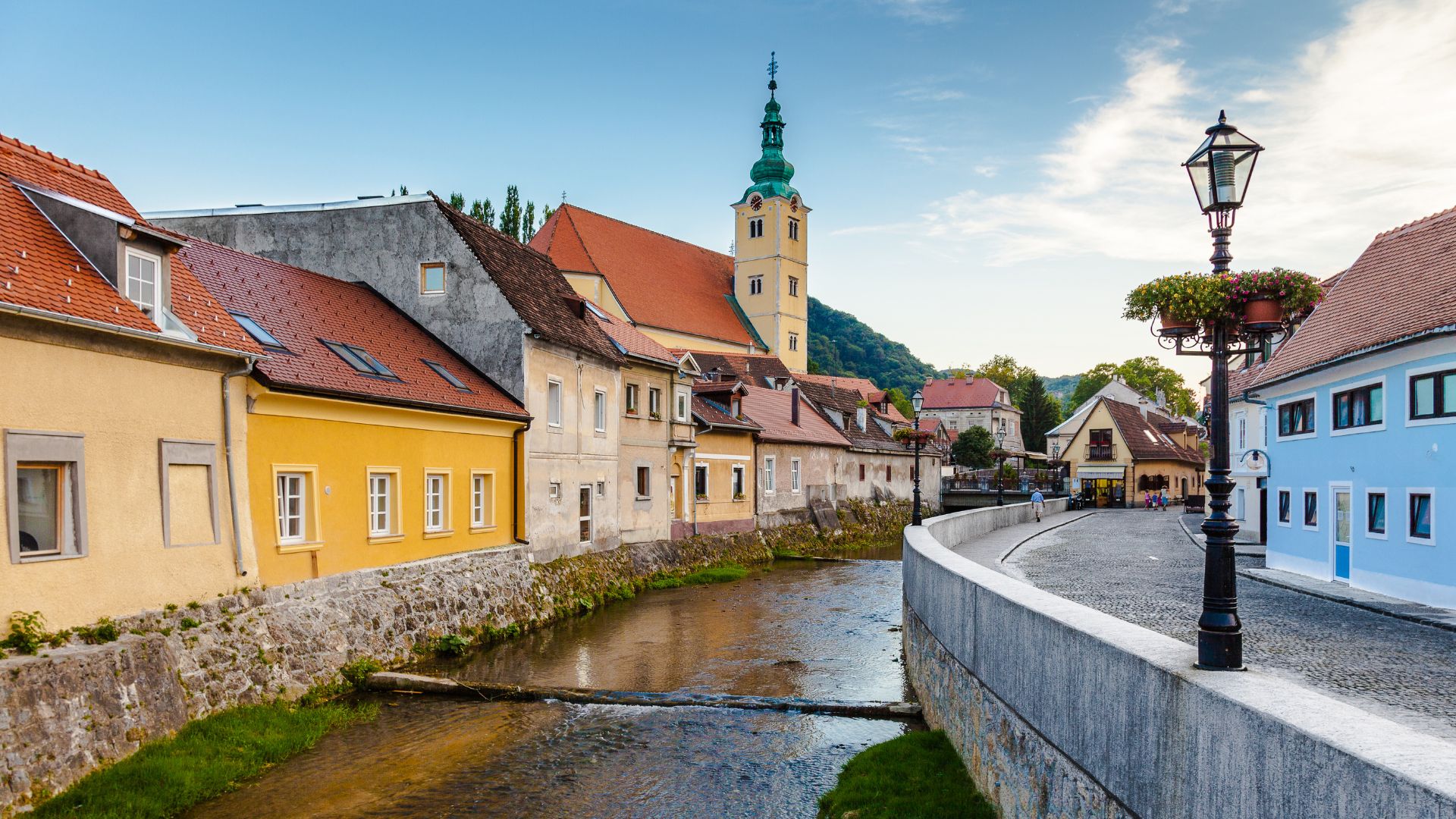 A picturesque view of the Gradna River flowing through the charming town of Samobor, Croatia, with colorful buildings lining the riverbank and a prominent church tower in the background, all under a bright sky.