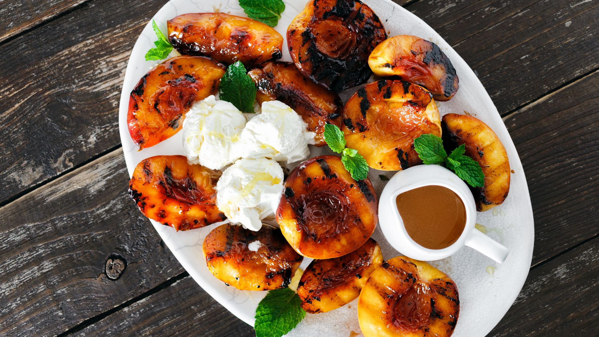 A white platter filled with golden-brown grilled peach halves, some drizzled with a dark, glossy sauce and served alongside scoops of white ice cream or whipped cream, garnished with fresh mint leaves, all resting on a rustic wooden table.