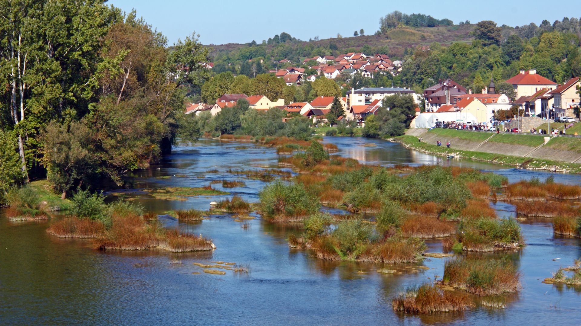 A wide shot of a tranquil river with numerous small, vegetated islands, leading towards a town with houses nestled among trees on the riverbank and a hilly background under a clear sky.