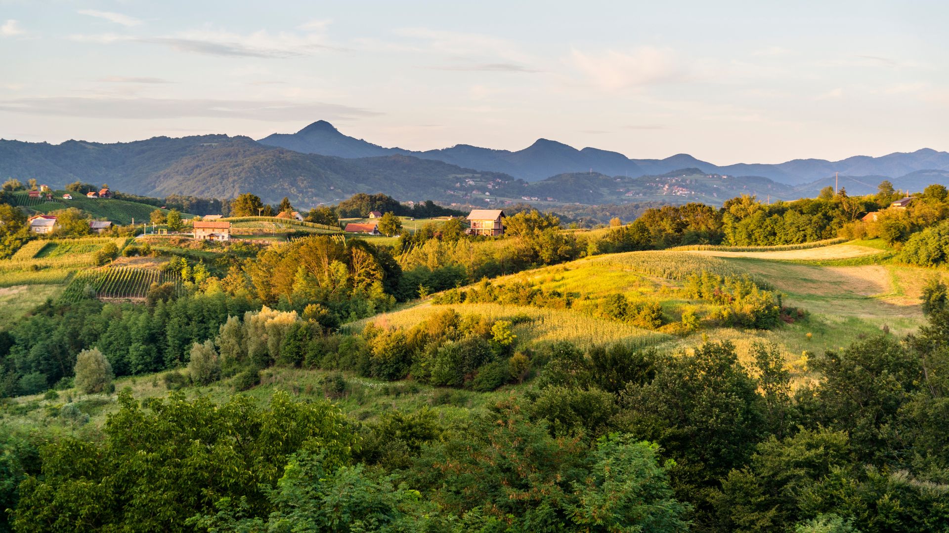 A panoramic view of rolling green hills and valleys in a rural European landscape, with scattered houses nestled among trees and fields, and distant mountains under a bright sky.