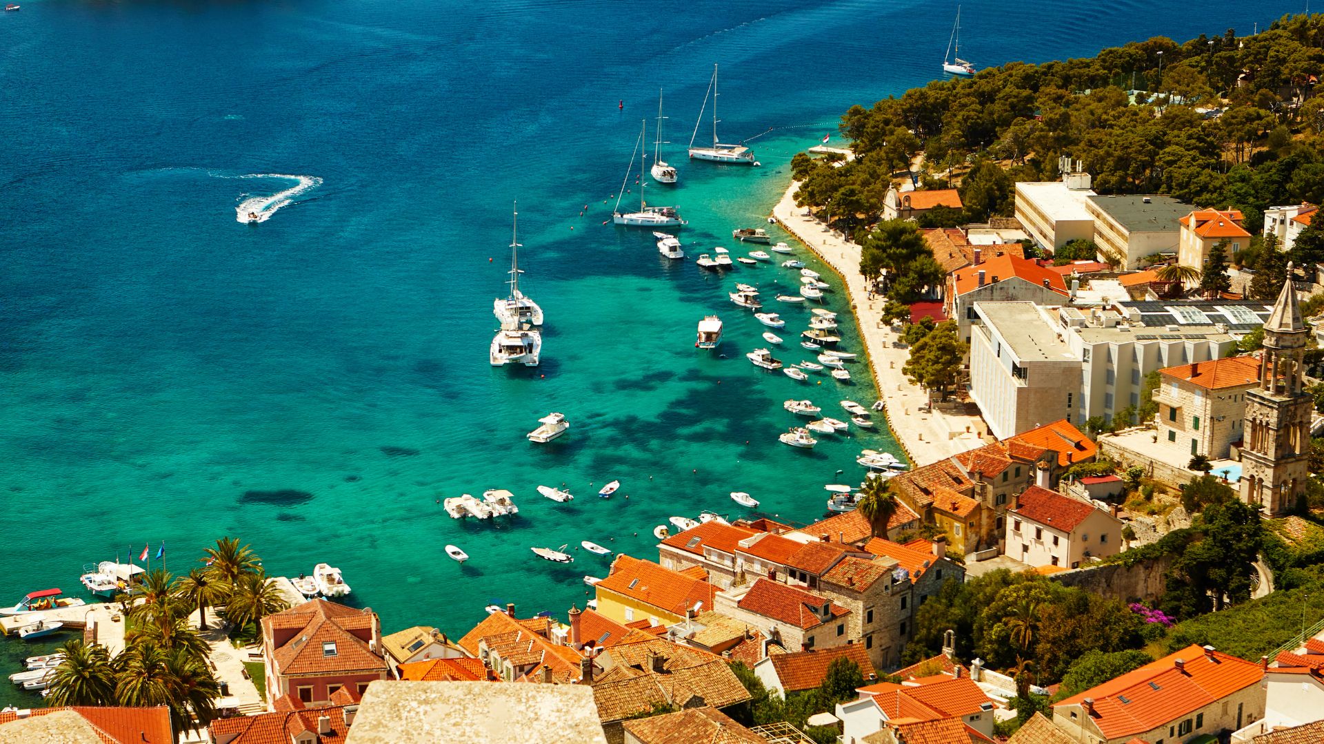 A vibrant aerial view of Hvar Town, Croatia, showing terracotta-roofed buildings clustered along a turquoise bay filled with numerous boats and yachts, with lush green hills rising behind the town. 