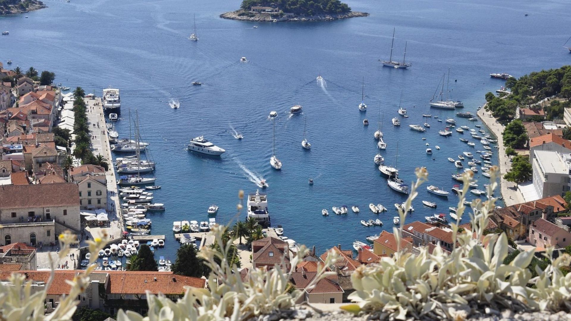 An aerial view showcases the bustling harbor of Hvar, Croatia, with numerous boats and yachts dotting the clear blue waters, surrounded by the town's buildings with terracotta roofs and lush green islands in the distance under a bright sky. 