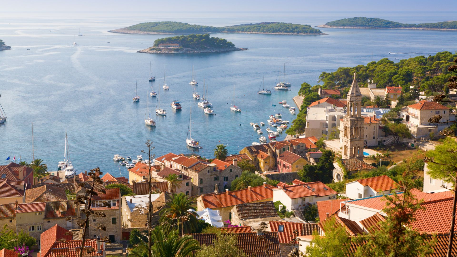A panoramic view of Hvar Town in Croatia, featuring red-tiled roofs, a prominent church tower, and a harbor filled with boats, surrounded by the clear blue waters of the Adriatic Sea and several small islands in the distance.