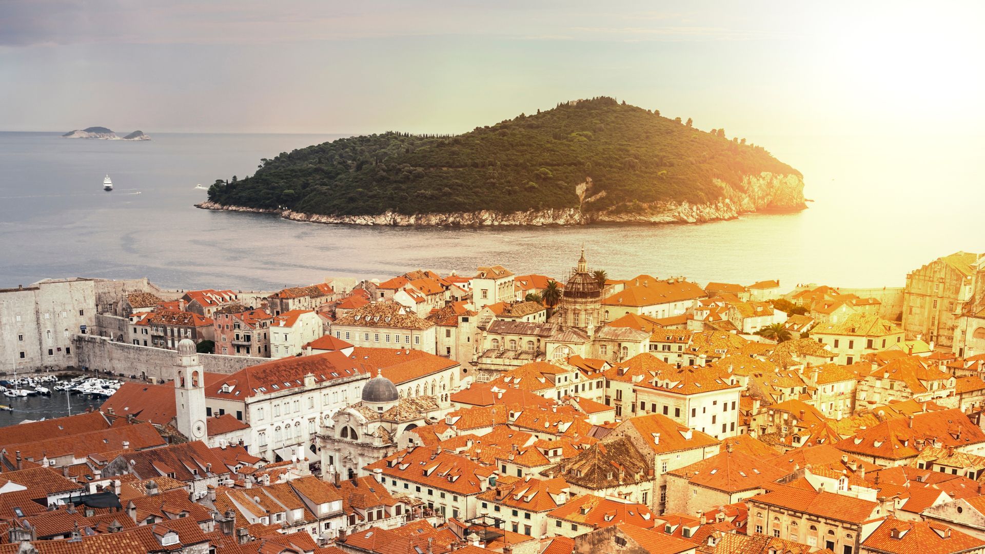 A panoramic view of Dubrovnik's red-roofed Old Town nestled along the coast, with the lush green Lokrum Island situated in the calm Adriatic Sea under a bright sky with a visible sun flare on the right.