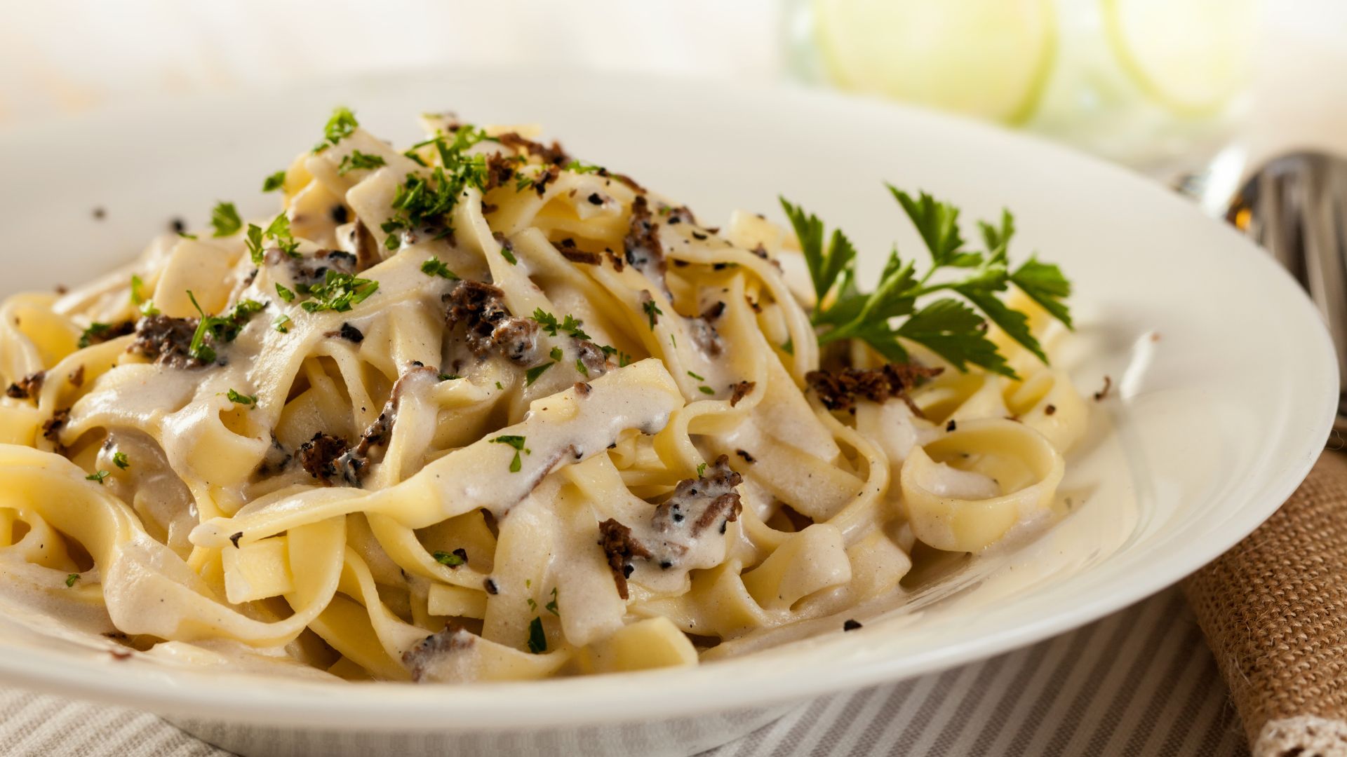A close-up shot of a plate of tagliatelle pasta with a creamy sauce, garnished with dark truffle shavings and fresh parsley.