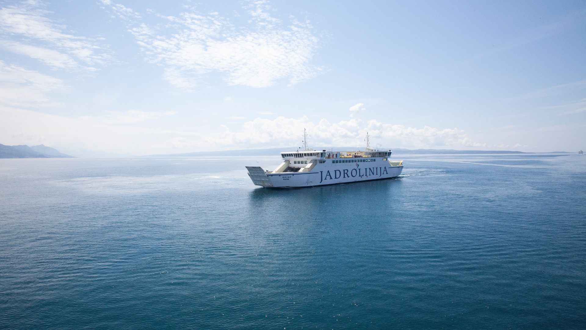 A large white Jadrolinija car ferry sails across a calm, deep blue sea under a bright, partly cloudy sky, with distant land visible on the horizon.