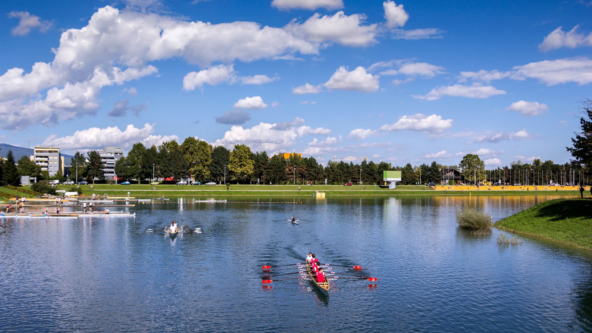 A panoramic view of Jarun Lake in Zagreb, Croatia, on a sunny day with a blue sky and white clouds. Several rowing boats are visible on the calm water, with people participating in rowing activities. The lake is surrounded by green banks, trees, and distant buildings under a bright sky, suggesting a recreational area. 