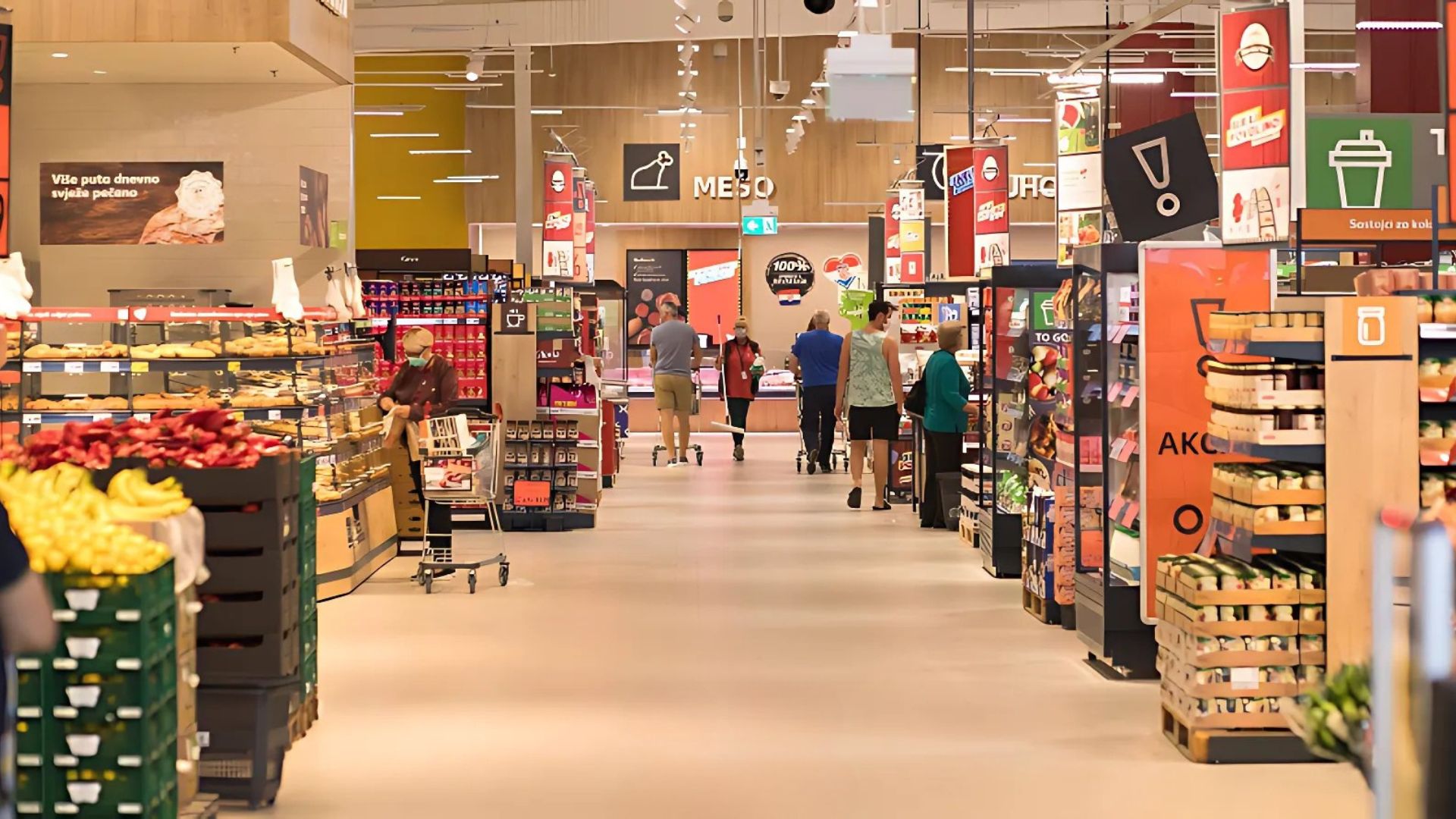 An indoor view of a brightly lit supermarket aisle, likely a Kaufland store, with shelves stocked with various products, including baked goods, produce, and packaged items. Several shoppers are visible in the distance, and signage in a Slavic language is displayed throughout the store.