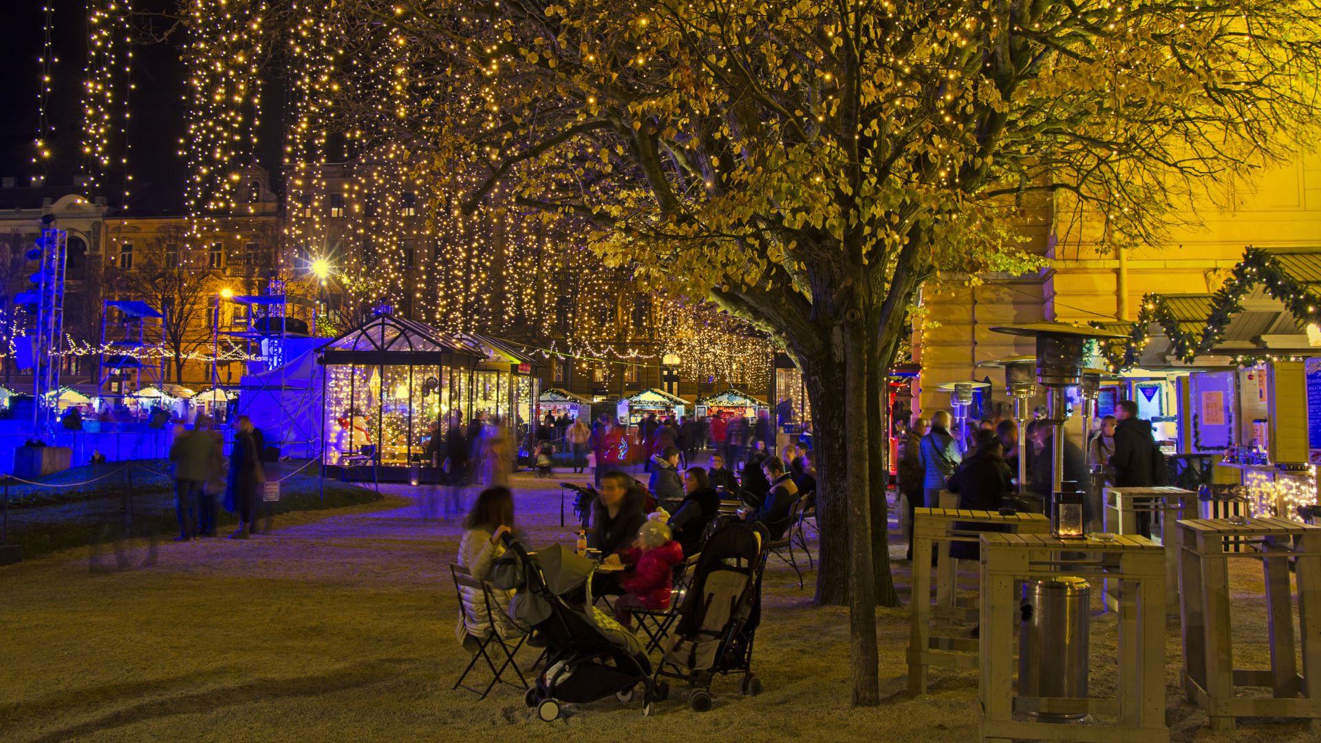 A night scene of a lively European square, possibly a Christmas market, illuminated by numerous warm string lights hanging from above and strung through the trees, with people dining at outdoor tables and strolling through the festive area.