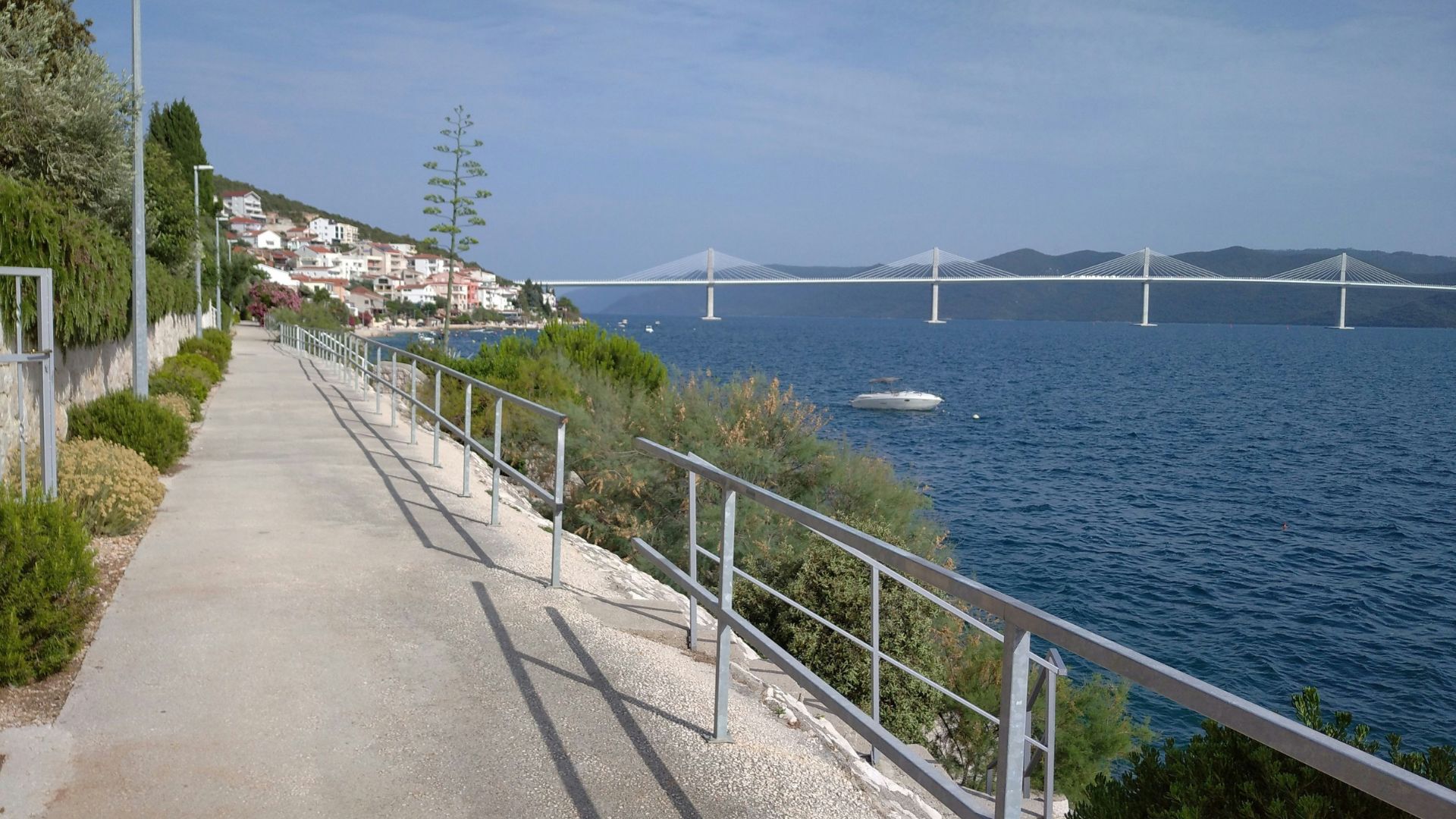 A paved waterfront promenade with a silver railing on the right, overlooking a calm blue sea with a white boat. In the distance, a modern cable-stayed bridge spans the water, connecting landmasses with a town visible on the left hillside.