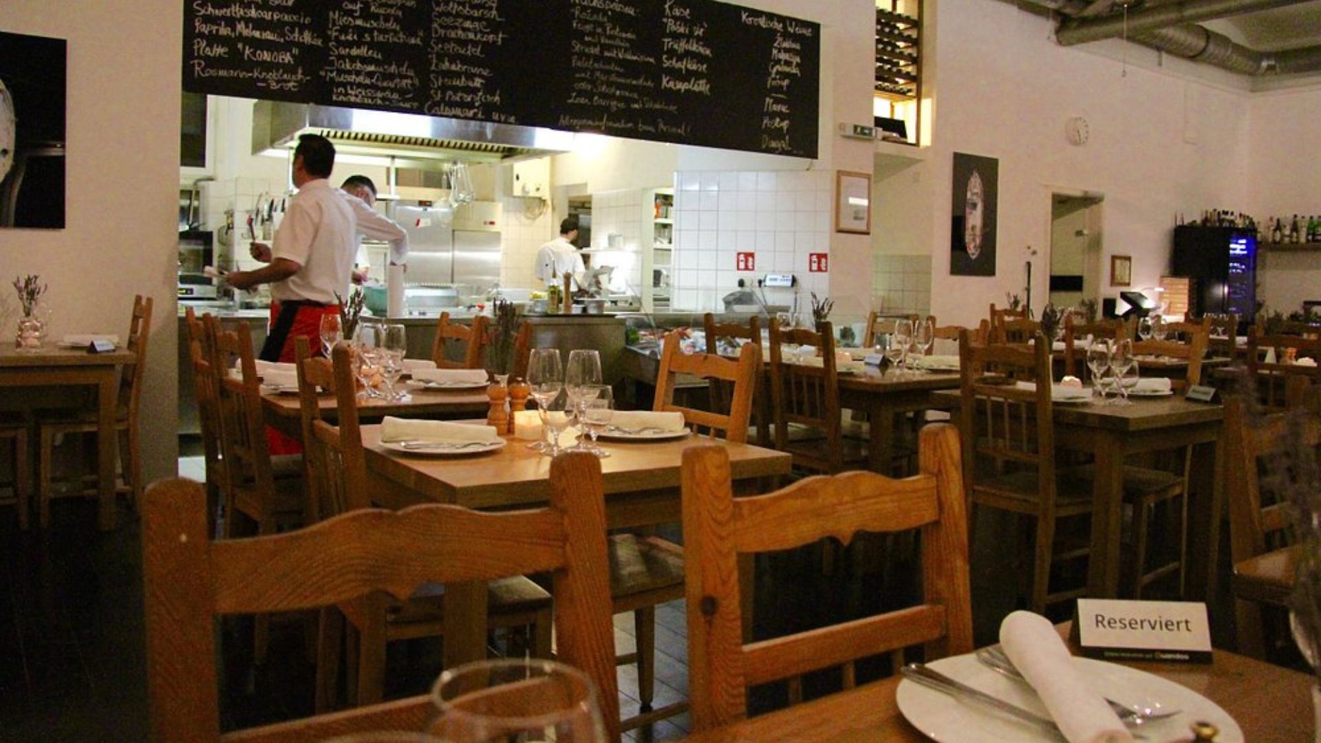 An interior shot of a rustic restaurant with wooden tables and chairs, an open kitchen in the background where chefs are working, and a large blackboard displaying a menu in what appears to be Croatian, with "KONOBA" visible as a menu item. A "Reserviert" (Reserved) sign is visible on a table in the foreground.