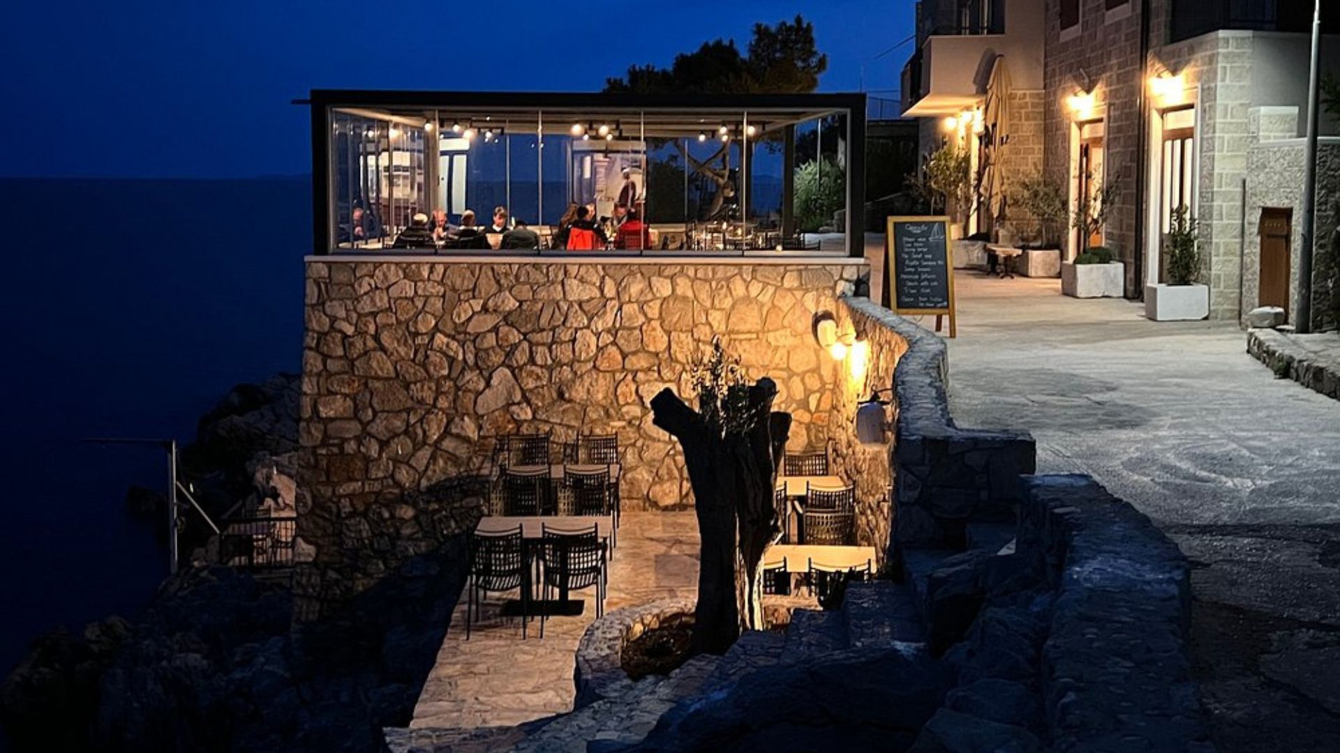 A nighttime view of a stone building with a glass-enclosed dining area overlooking the sea, and an outdoor patio with tables and chairs in the foreground, illuminated by warm lights.