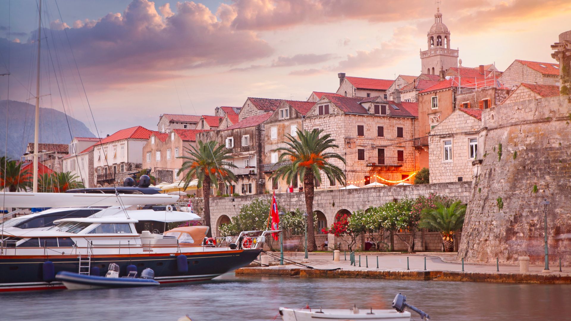 A scenic view of Korčula's Old Town in Croatia, featuring historic stone buildings with red-tiled roofs, a prominent church tower, and palm trees along a waterfront where boats and yachts are docked in calm, clear waters under a vibrant sunset sky.