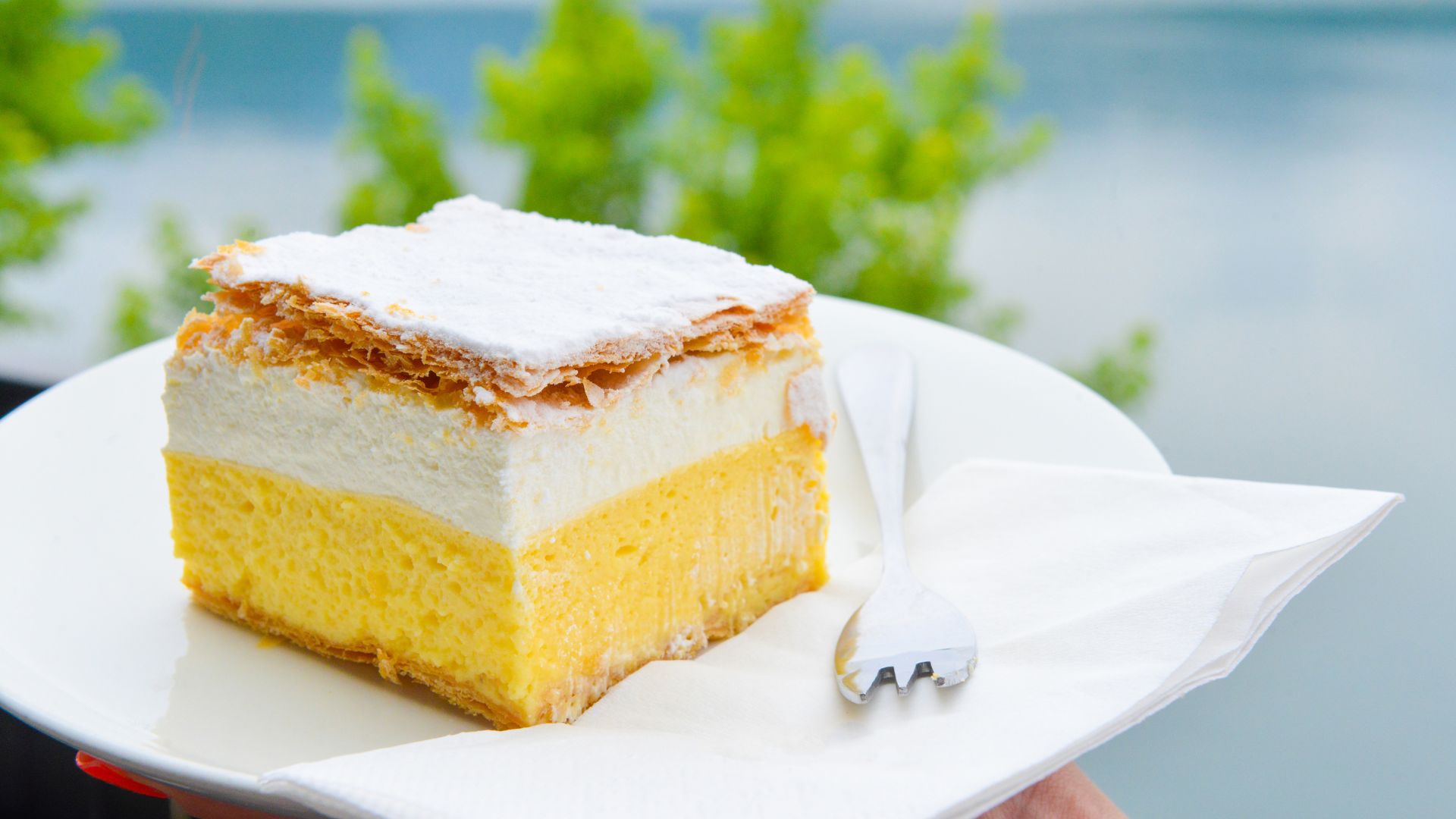 A close-up shot of a slice of Kremšnita (cream cake) on a white plate, featuring layers of puff pastry, vanilla custard, and whipped cream, topped with powdered sugar, with a white fork resting on a napkin beside it.
