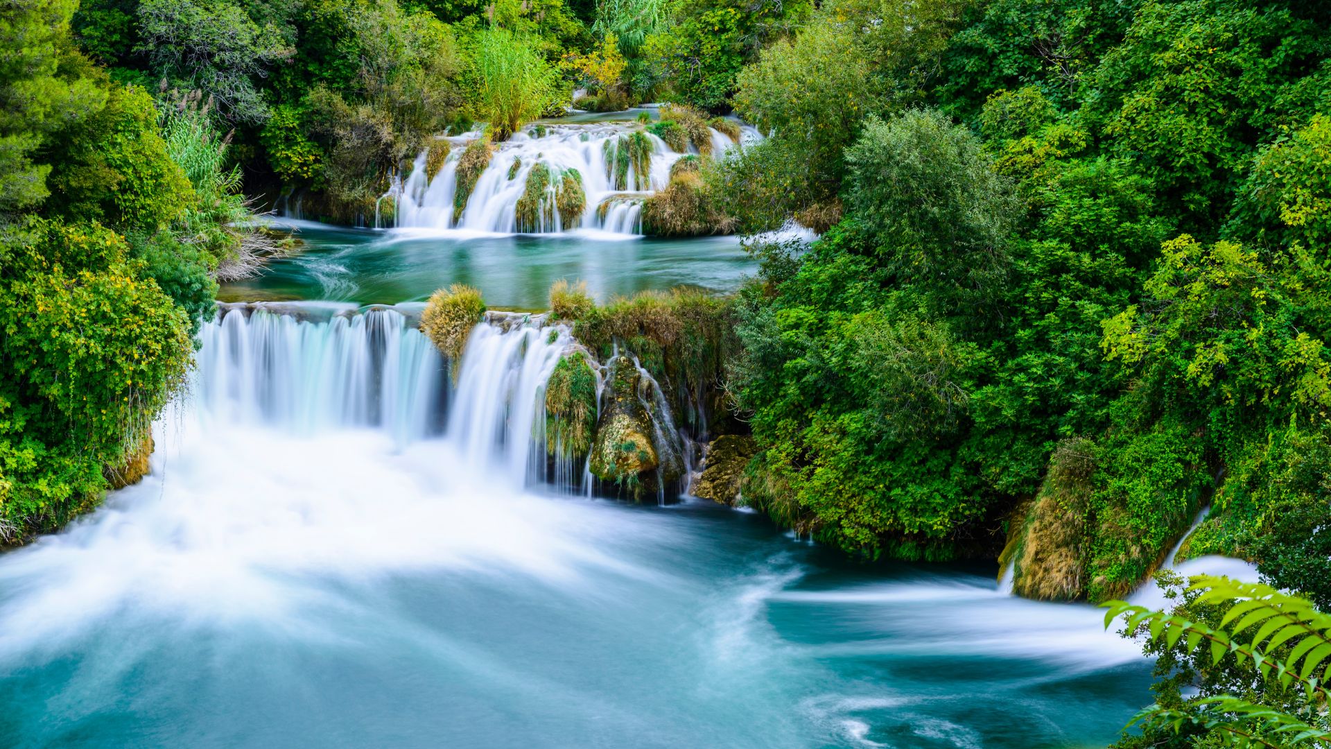 A vibrant, long-exposure photograph of the multi-tiered waterfalls at Krka National Park in Croatia, showing turquoise water cascading over travertine formations surrounded by lush green foliage.