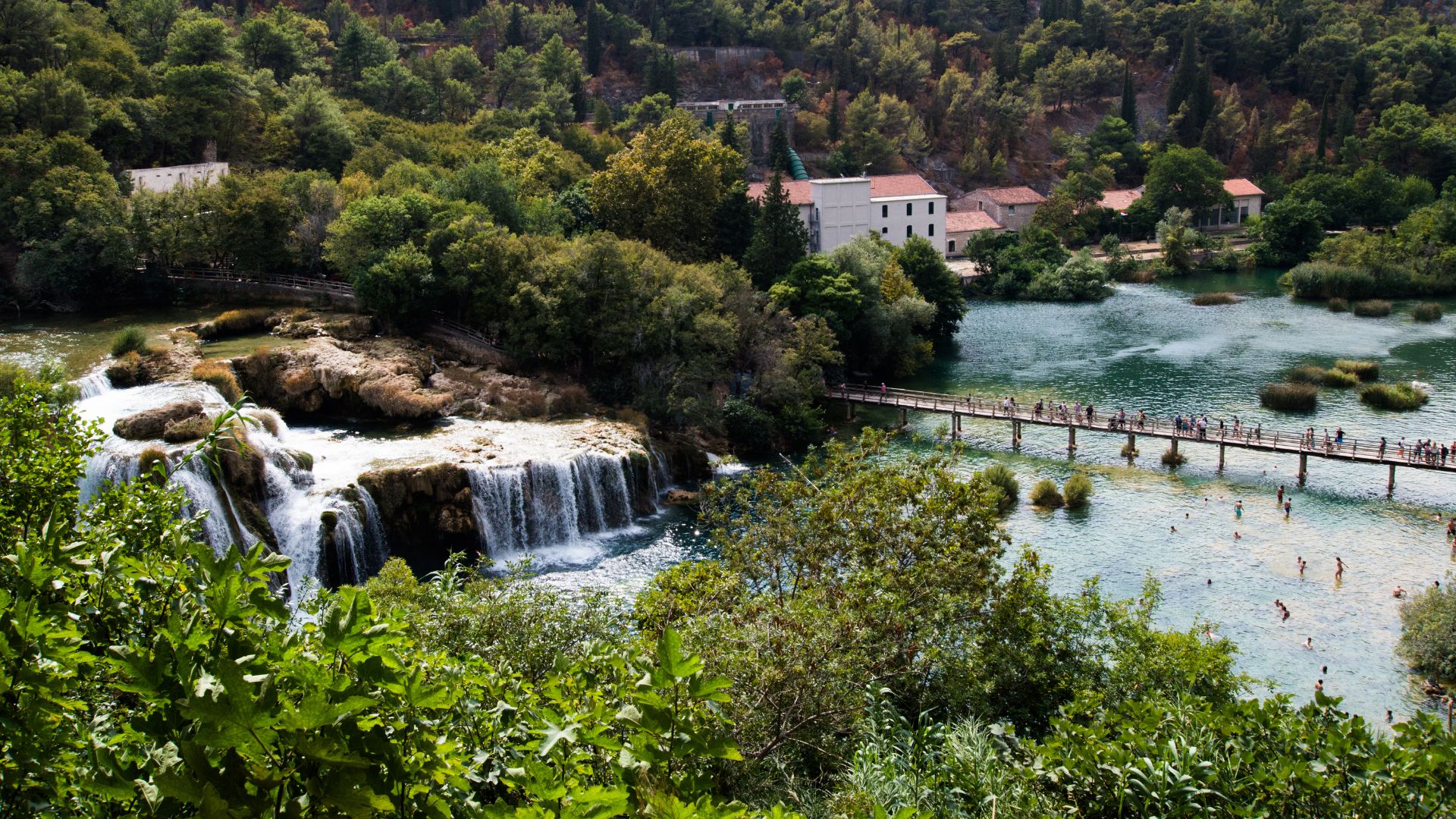 A wide shot of the Skradinski Buk waterfall in Krka National Park, Croatia, showing multiple cascades flowing into a clear river where people are swimming, with a long wooden bridge spanning the river and lush green foliage and buildings in the background.