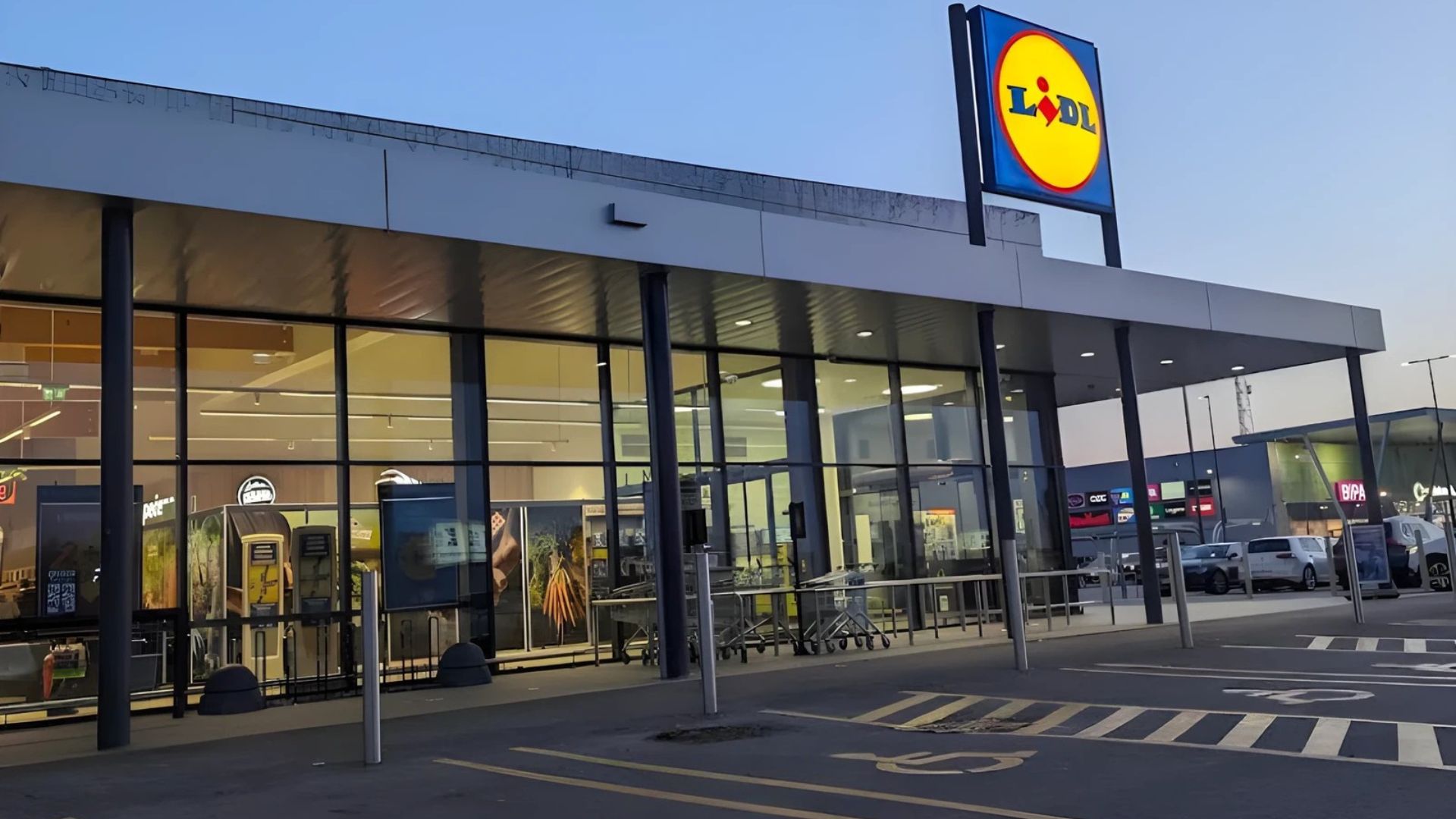 This image shows the exterior of a Lidl supermarket, a large, modern building with a prominent yellow and blue Lidl logo sign above the entrance. The store features a wide glass facade, allowing a view into the interior, and a parking lot with marked spaces in the foreground. 