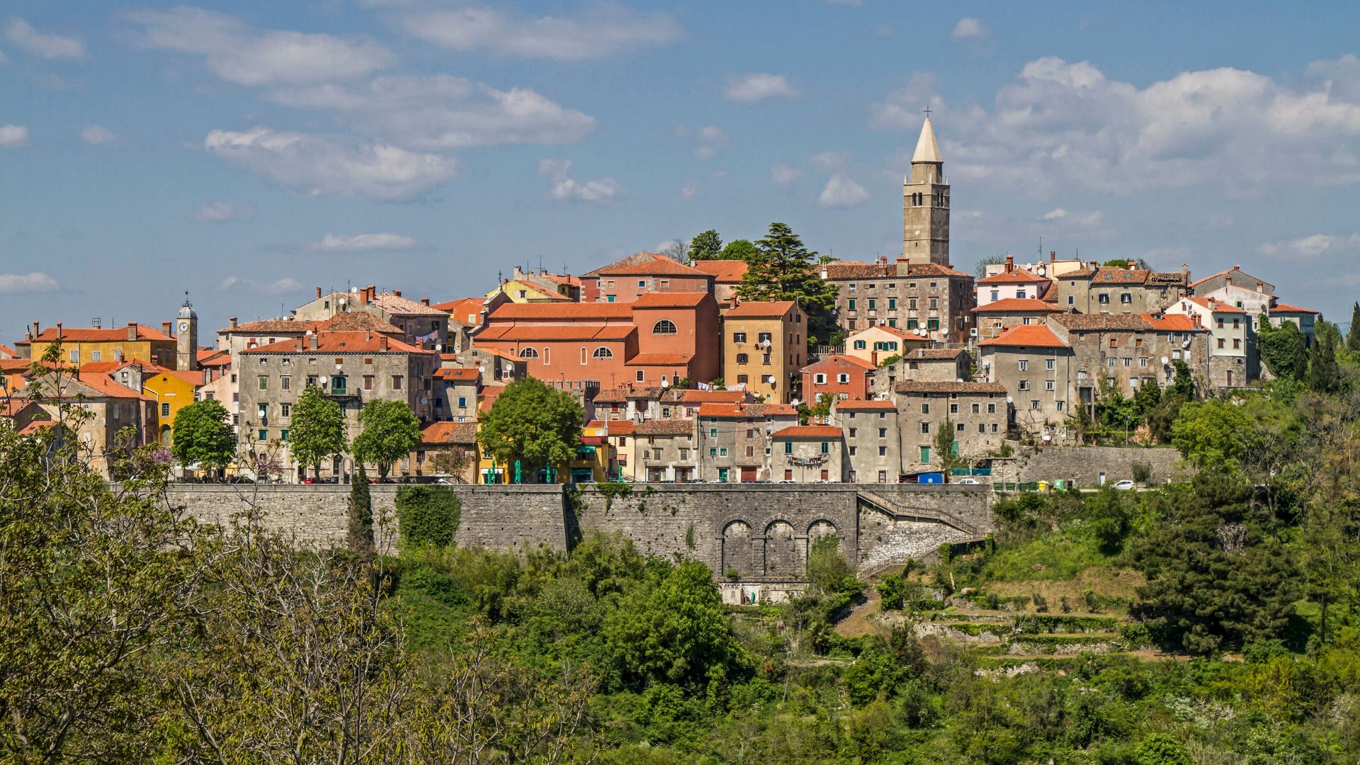 A picturesque medieval town with red-tiled roofs and stone buildings, dominated by a tall bell tower, situated on a hill overlooking lush green landscapes under a partly cloudy sky.