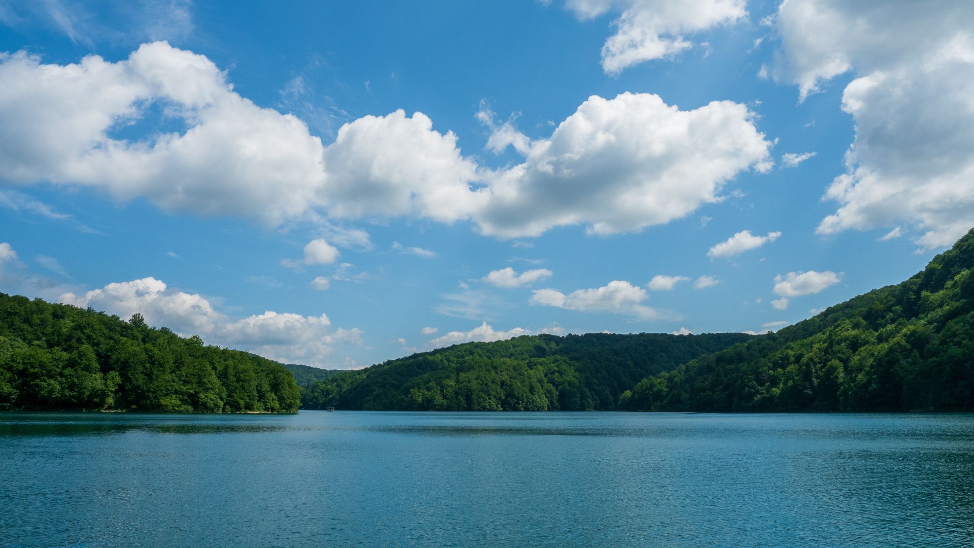 A serene wide shot of a large, turquoise lake surrounded by lush green forested hills under a bright blue sky with scattered white clouds.