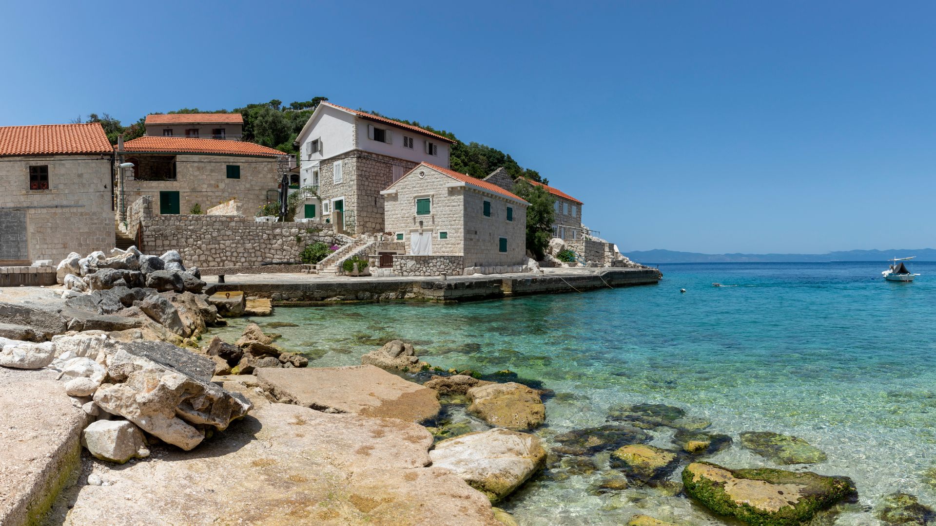 A coastal village with stone houses and red-tiled roofs situated along a rocky shoreline next to clear turquoise waters, with a small boat visible in the distance under a bright blue sky.