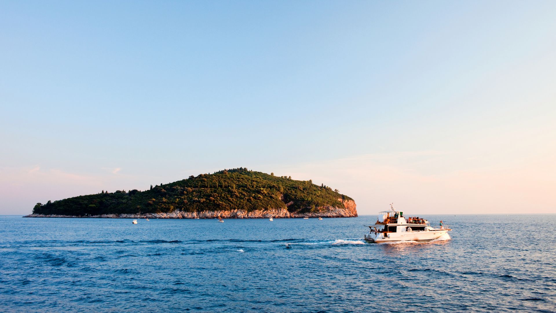 A small, lush green island covered in trees sits in the clear blue sea under a bright sky, with a white boat moving across the water in the foreground, leaving a wake behind it.