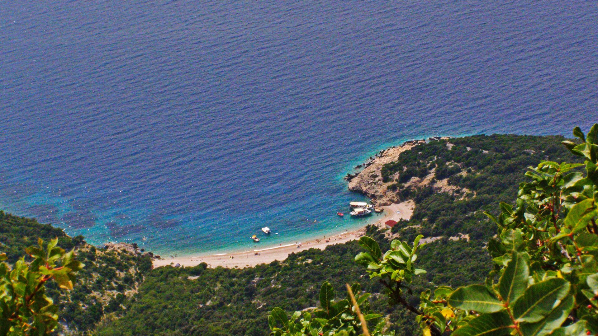 An aerial view shows a secluded white pebbly beach nestled in a cove with turquoise waters, surrounded by lush green hills and cliffs, with boats visible in the clear blue sea.