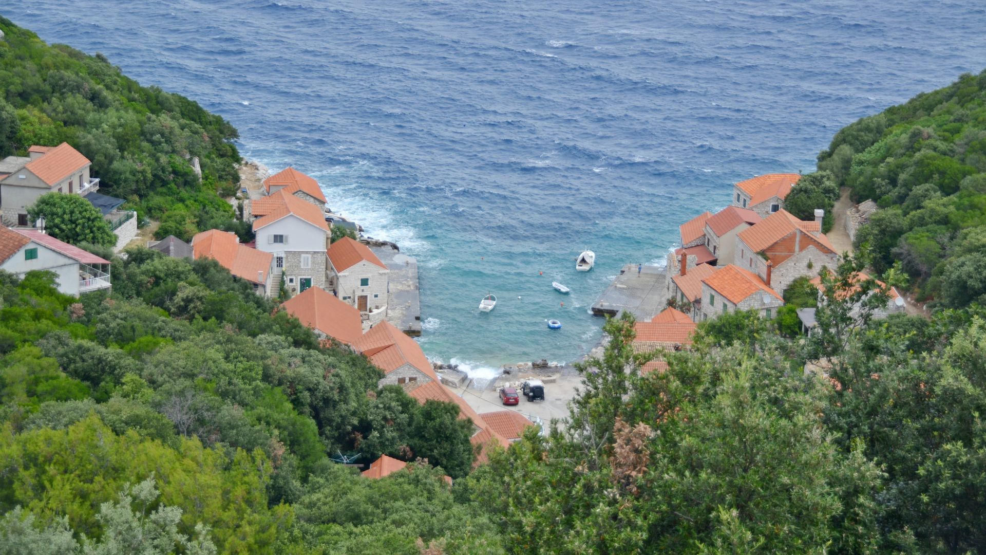 A high-angle view shows the scenic Lučica Bay in Lastovo, Croatia, featuring a cluster of traditional houses with red-tiled roofs nestled along the clear turquoise waters of a small bay, flanked by lush green hills, with several small boats anchored in the water.