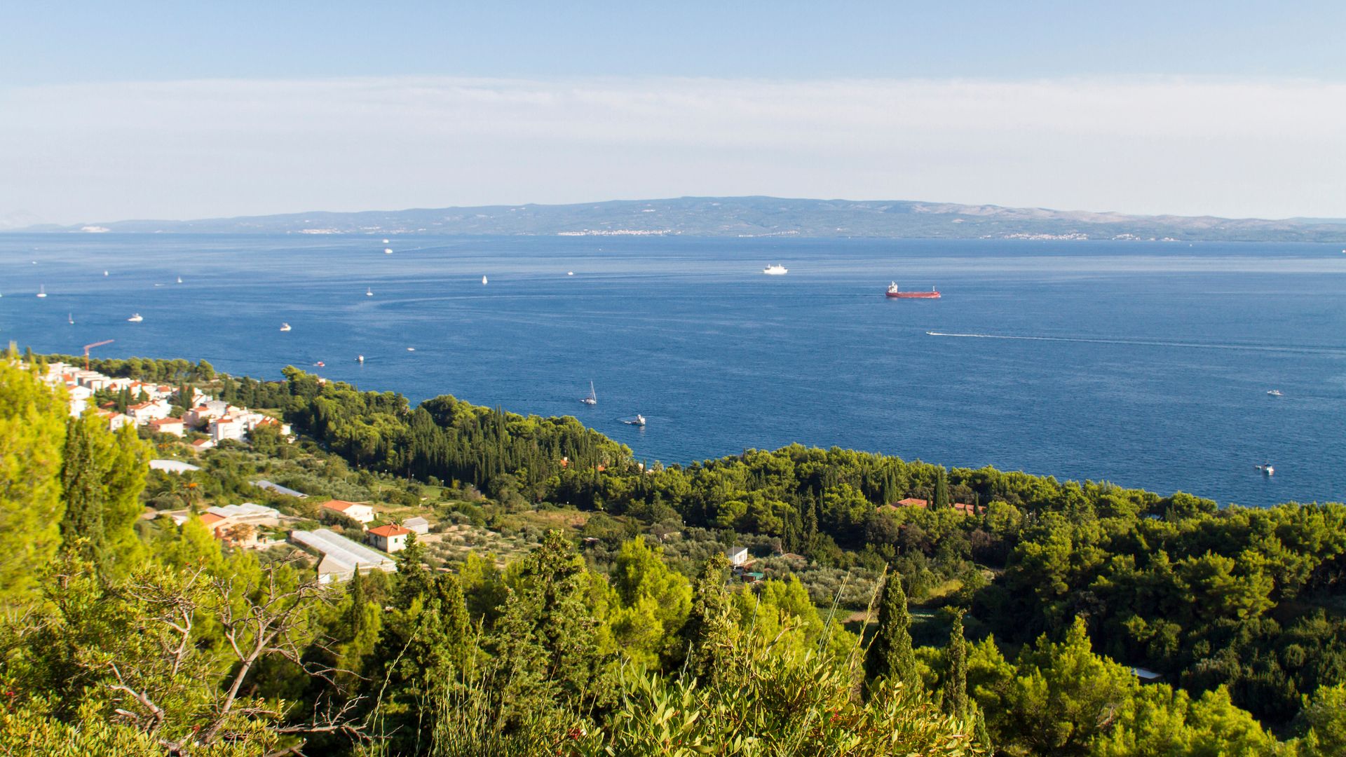 A panoramic view of the Adriatic Sea and the coastline of Split, Croatia, featuring a lush green hillside with pine forests, scattered buildings, and boats dotting the calm blue water under a clear sky.