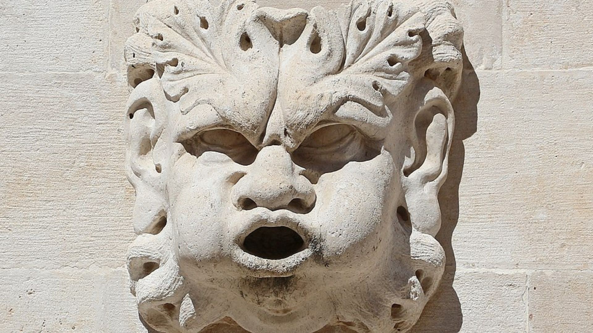 A close-up shot of a weathered, light-colored stone carving of a grotesque, expressive face with leaf-like elements around the forehead and ears, set against a light stone wall, casting a shadow to its right.