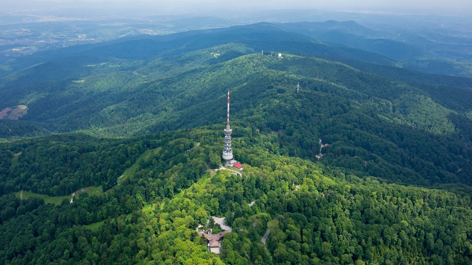 An aerial view of a densely forested mountain range, featuring a tall, slender communication tower on the central peak, with smaller structures and winding paths visible through the trees.