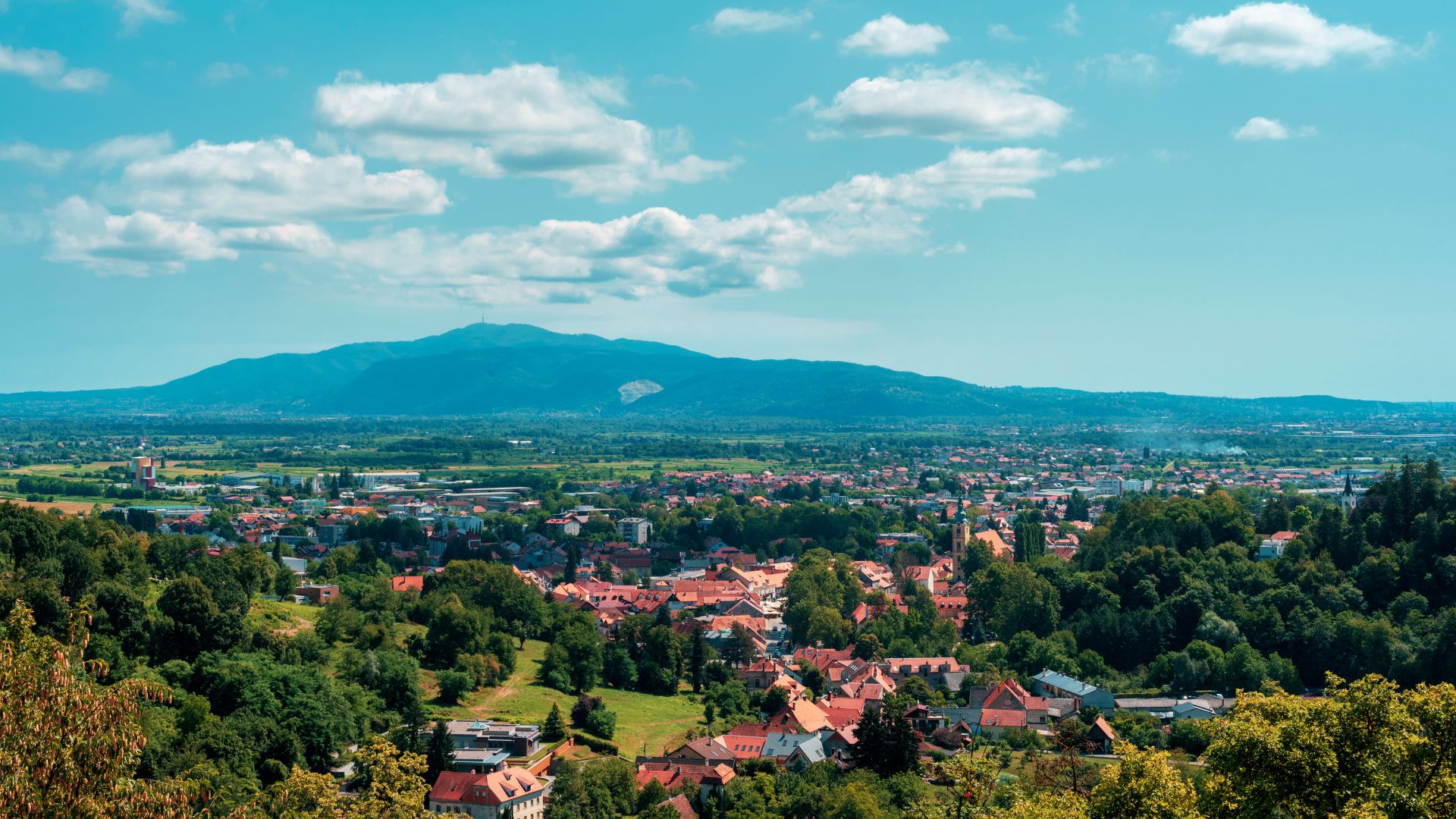 A panoramic view of a town with red-roofed buildings and lush green trees, set against the backdrop of a large mountain range under a blue sky with scattered white clouds.