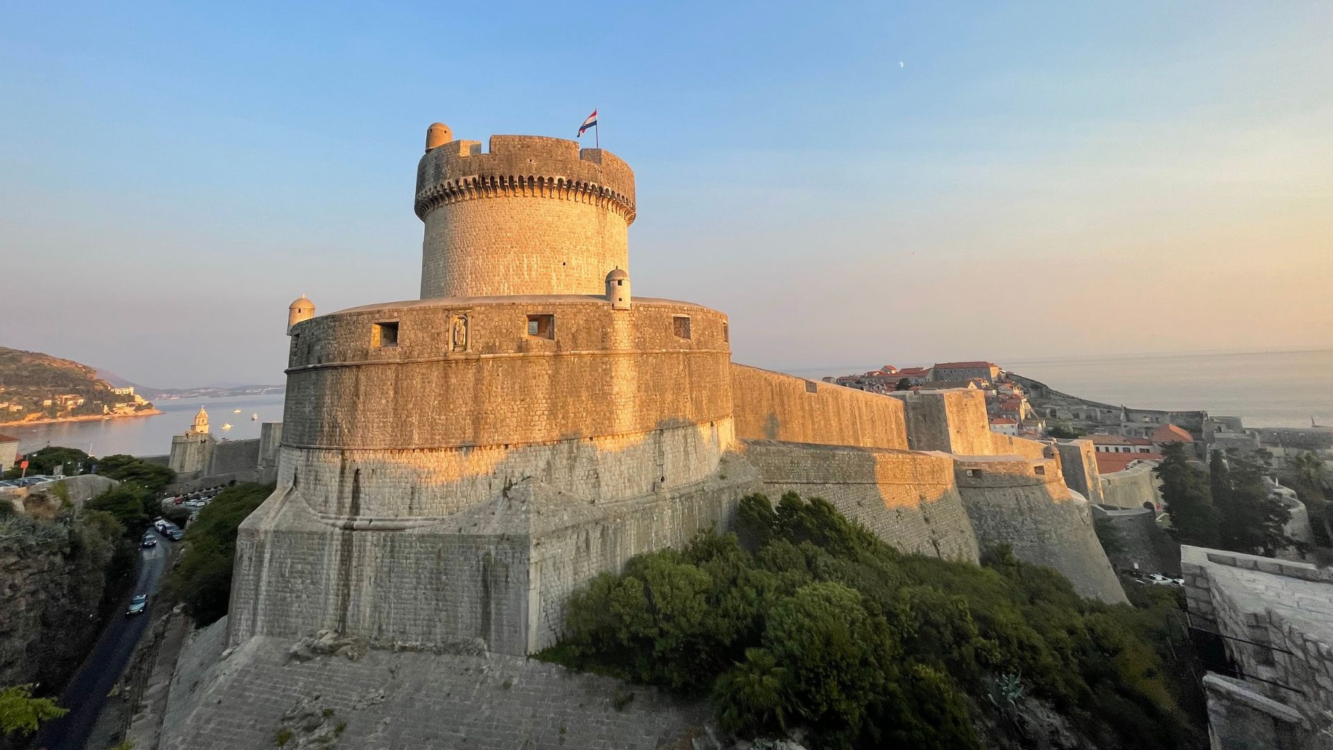 The image shows Minčeta Tower, a prominent circular stone fortress with a crenellated top, situated on a hill overlooking the historic city of Dubrovnik, Croatia, with the Adriatic Sea visible in the background under a clear sky during what appears to be sunrise or sunset. The tower is a key part of the city's defensive walls and offers panoramic views of the Old Town and the sea. 